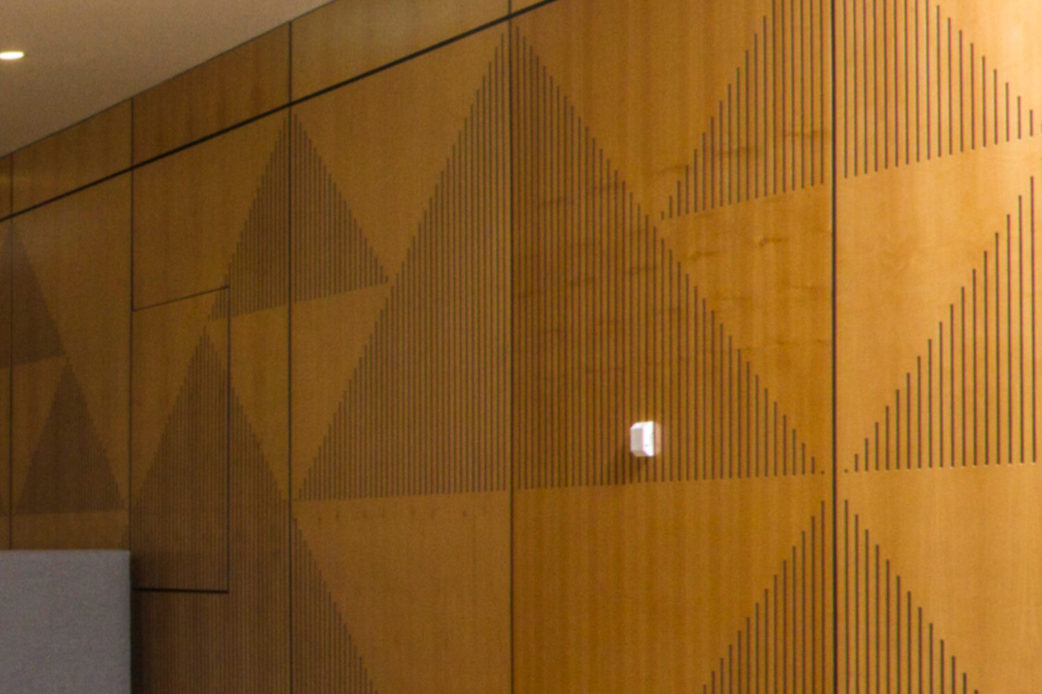 Wooden wall with geometric pattern made of thin black lines, with a small white device attached to the wall.