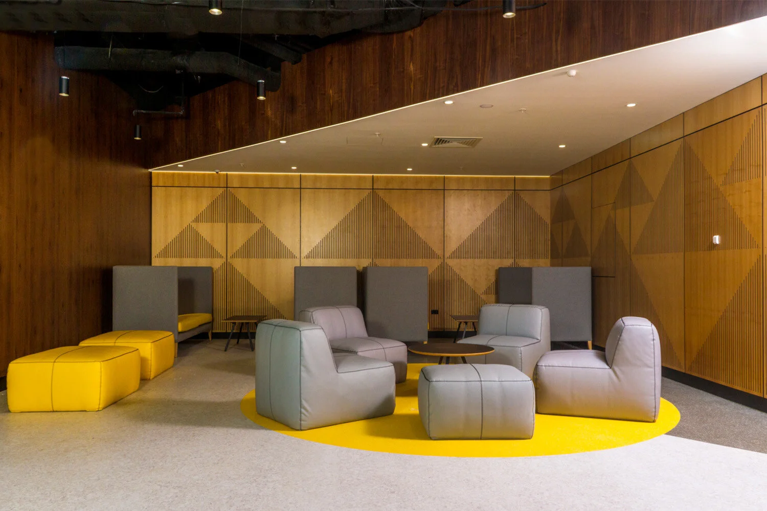 Modern lounge area featuring gray armchairs, yellow ottomans, and a yellow circular rug, with wooden wall panels and subdued lighting.