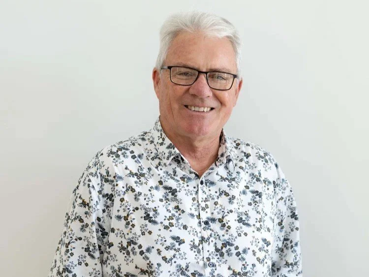 Man with white hair and glasses smiling, wearing a floral button-up shirt, standing against a plain white background.