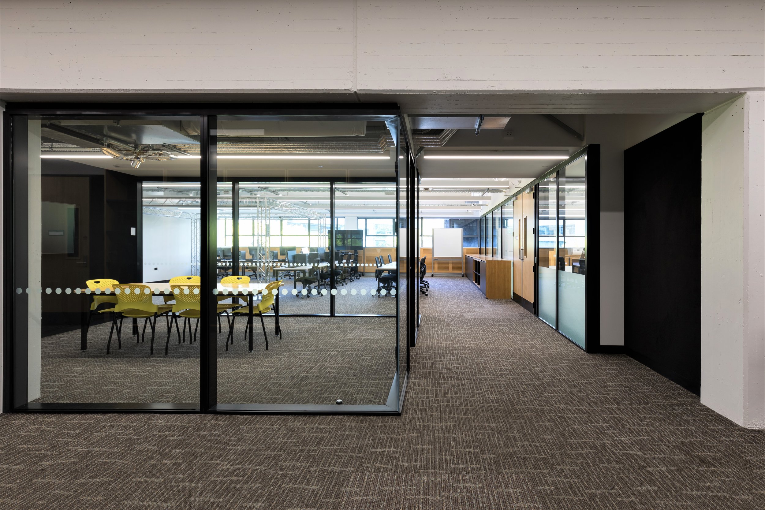 Modern office conference room with glass walls, yellow chairs around a table, and an open workspace area in the background.