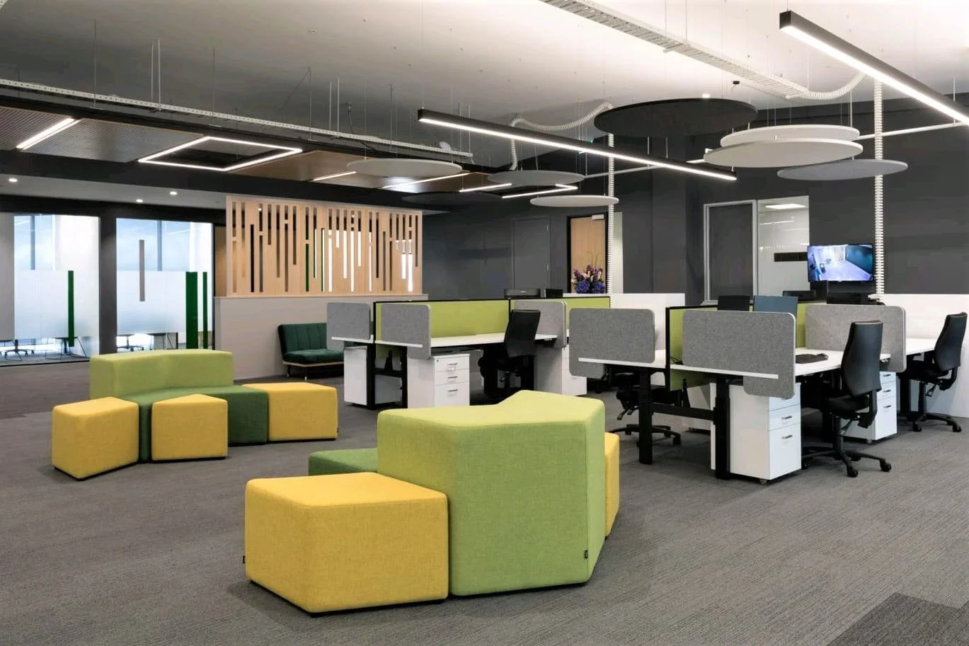 Modern open office space with green and yellow seating, workstations with partition panels, black office chairs, and ceiling lighting fixtures.