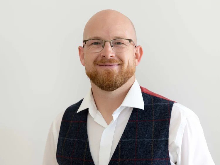 Portrait of a man with glasses, a beard, and a head shave, smiling, wearing a white shirt and a dark plaid vest, against a plain light background.