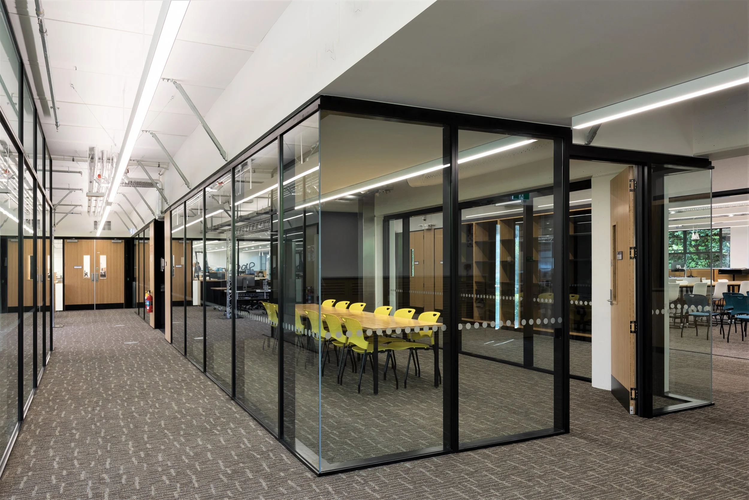 Modern office hallway with glass-walled conference room containing yellow chairs around a table.