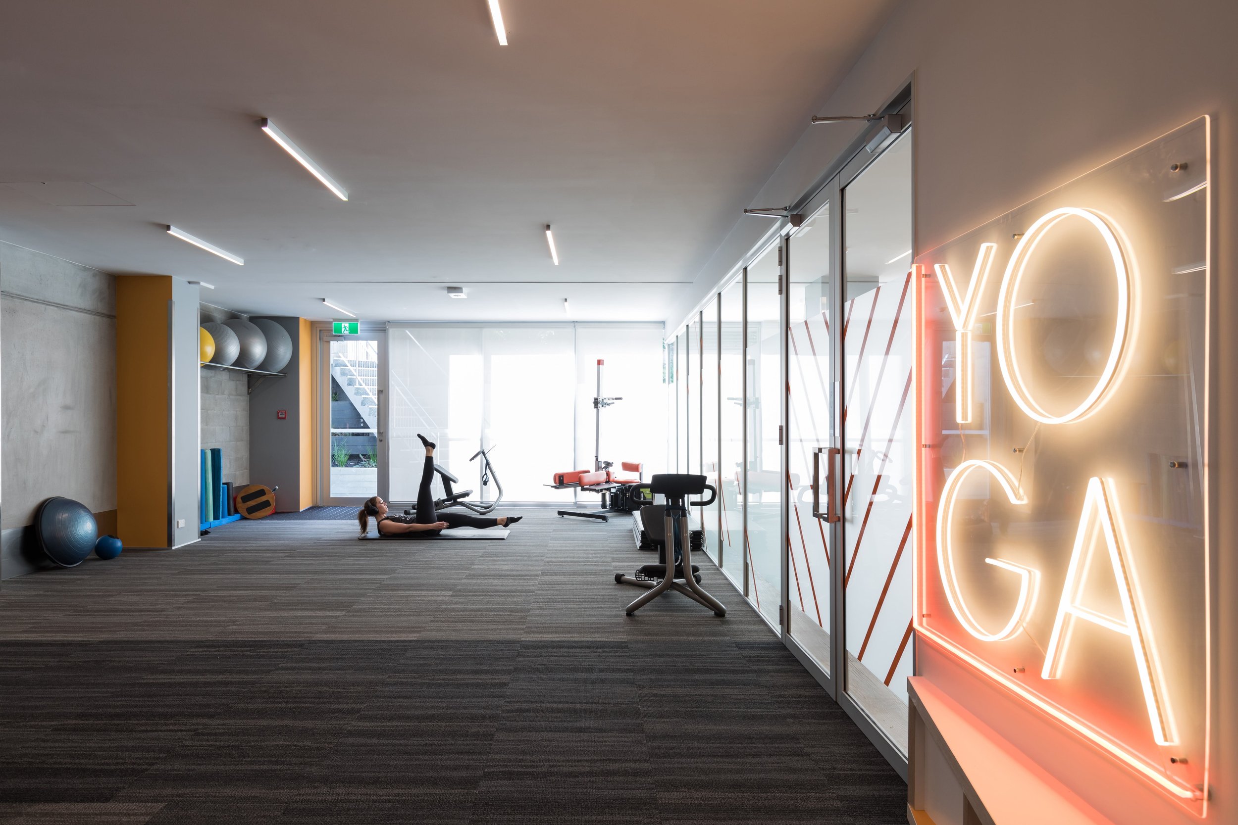 A woman lying on a workout mat doing leg lifts in a modern gym. The gym has exercise balls, mats, and equipment, with a large glass wall displaying neon lights spelling 'YOGA'.