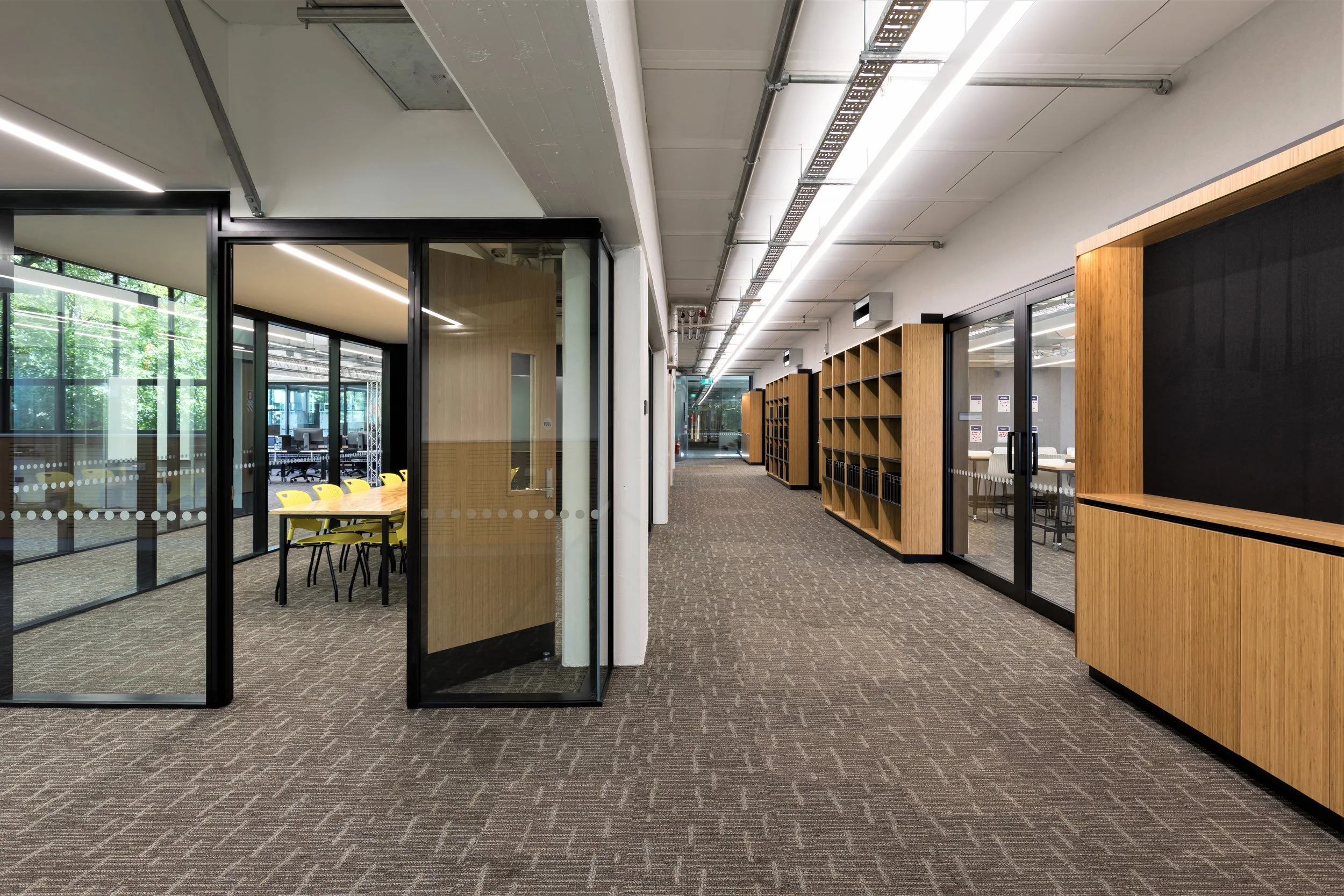 Modern office corridor with glass-walled conference rooms on the left, featuring a table and yellow chairs visible through the glass. On the right, wooden shelves and cabinets are along the wall. The corridor has brown patterned carpet, white walls, 