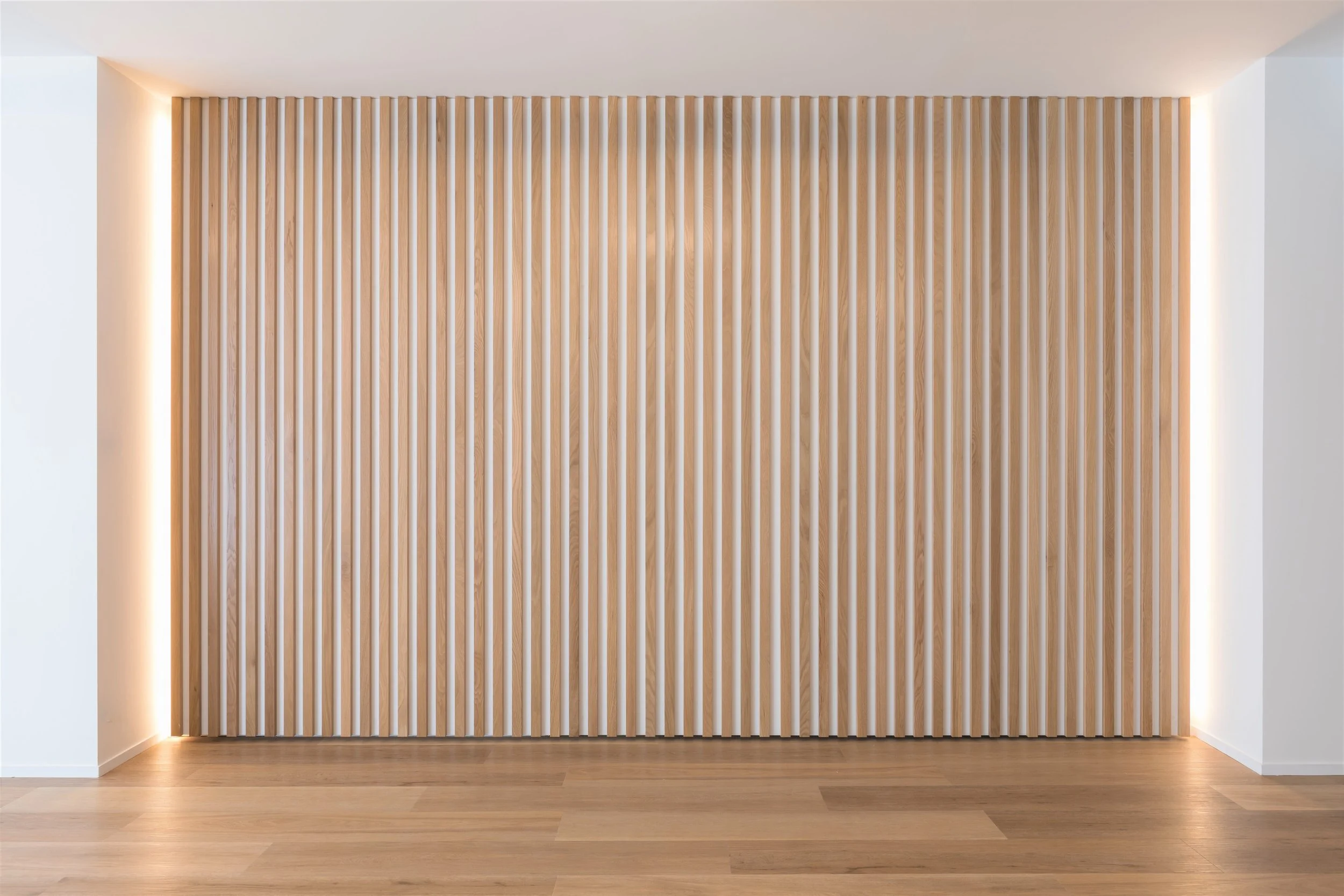 Modern interior wall with vertical wooden slats and hidden LED lighting along the top and bottom edges, wooden floor, white walls on either side.