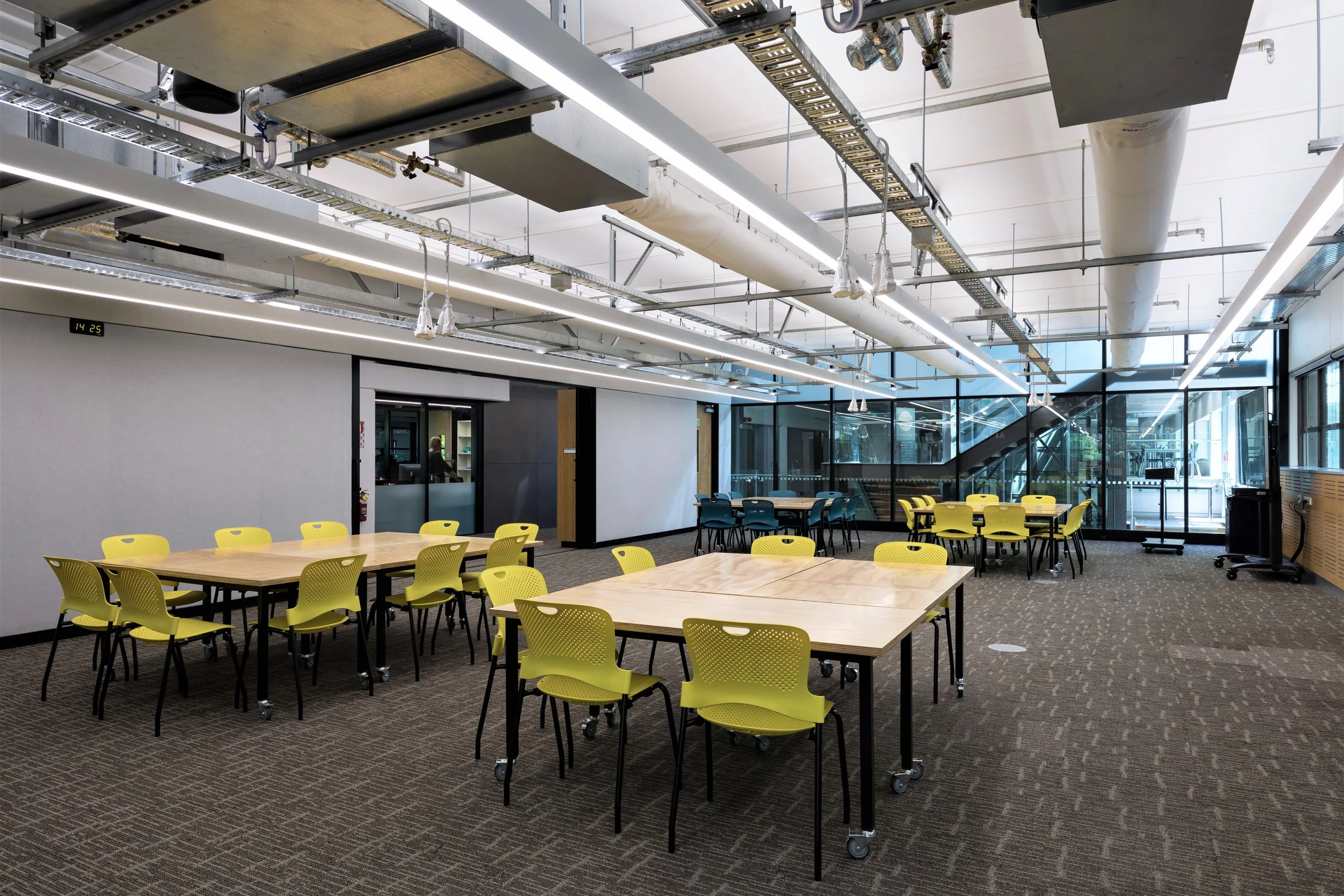 Empty modern conference room with yellow chairs and light wood tables, glass windows, and ceiling-mounted lighting fixtures.