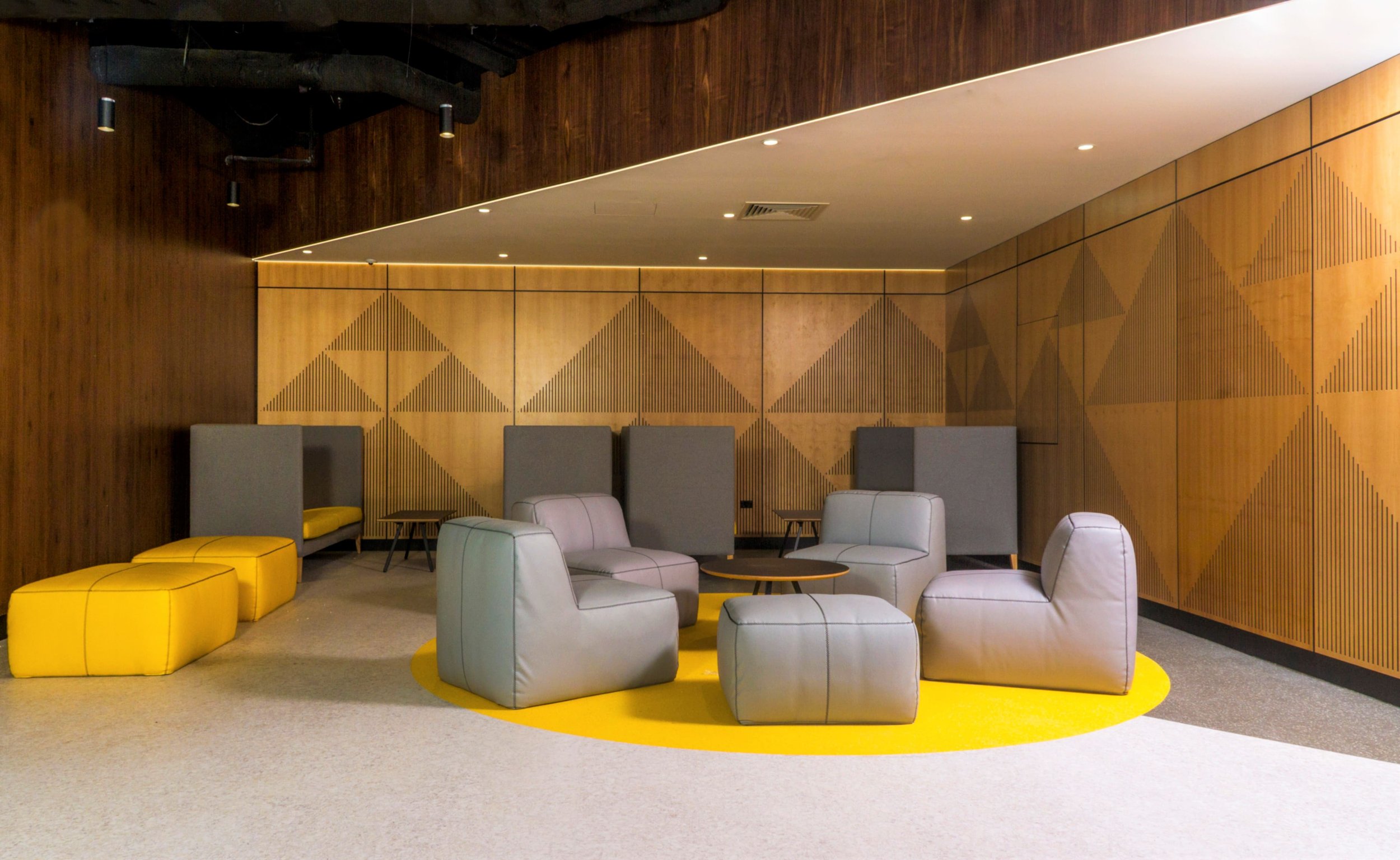 Modern lounge area with gray and yellow seating, wooden wall panels with geometric patterns, and small tables.