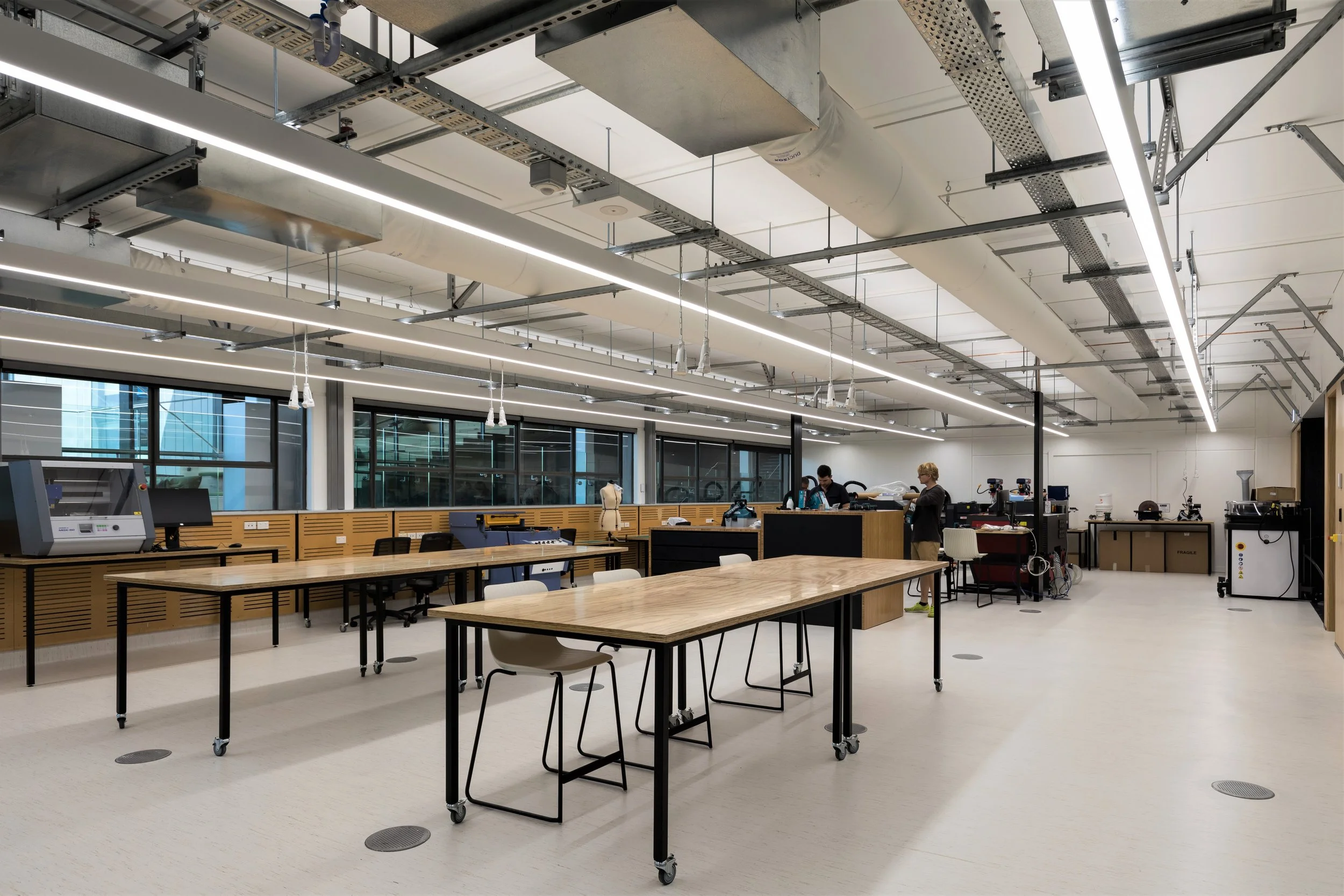 Empty laboratory or workshop with wooden tables, chairs, and scientific equipment, illuminated by overhead lights and large windows.