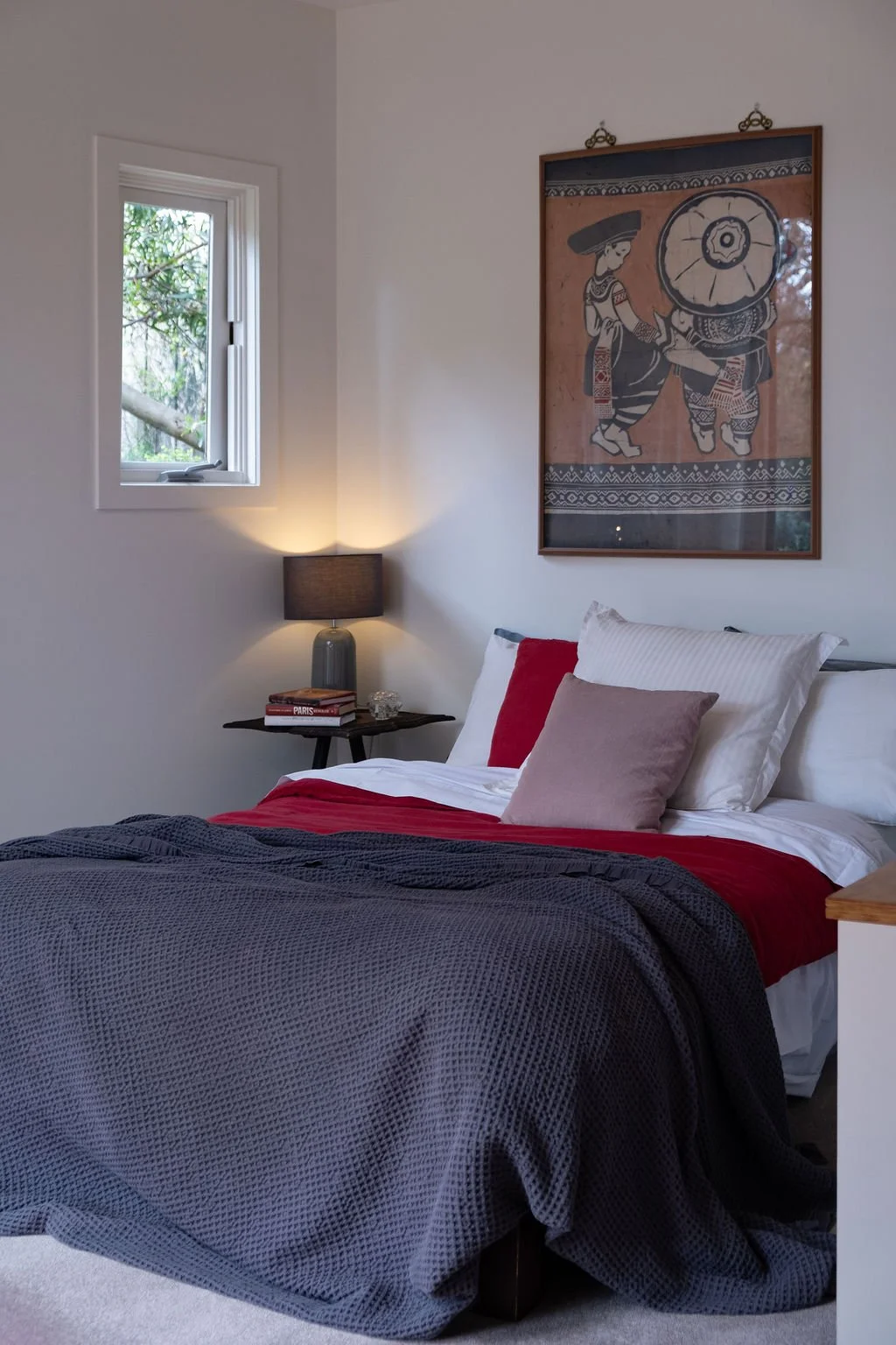 A bedroom with a bed that has white, gray, and red bedding and pillows, a nightstand with a black lamp and books, a window with greenery outside, and a framed artwork of traditional indigenous figures hanging above the bed.