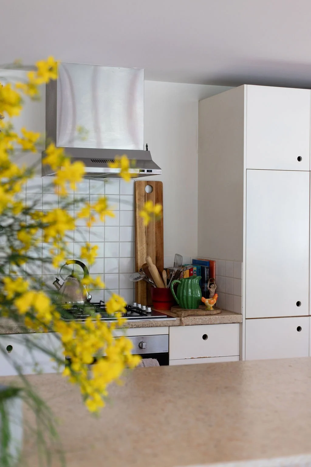 A cozy kitchen with white cabinets, a tiled backsplash, a wooden cutting board, a green pitcher, and a yellow flower in the foreground.