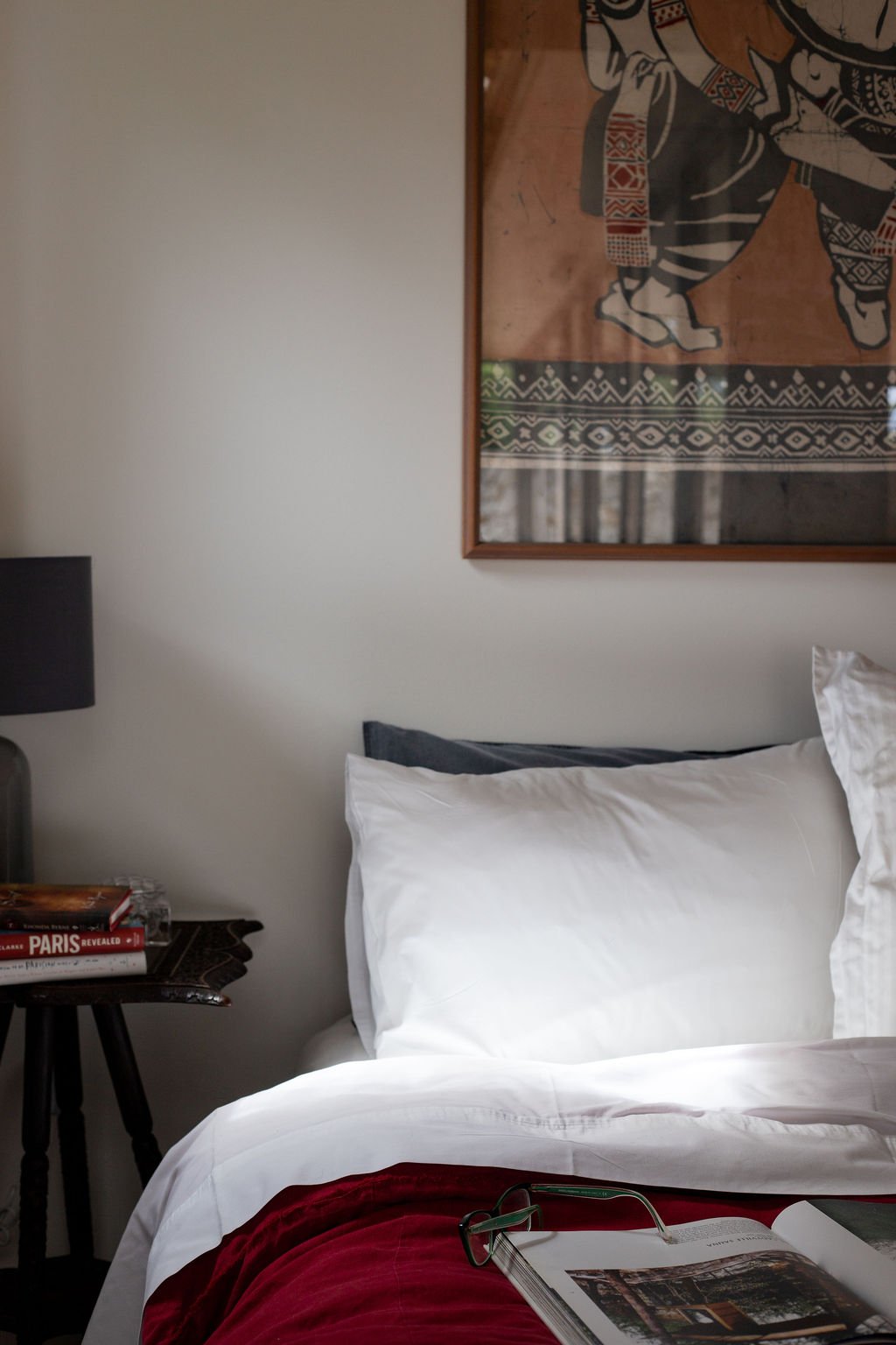 A bedroom with a bed covered in white linens, with a red throw blanket at the foot, and glasses resting on an open book. A small side table with books and a glass of water is next to the bed. There is a framed cloth or textile artwork hanging on the 