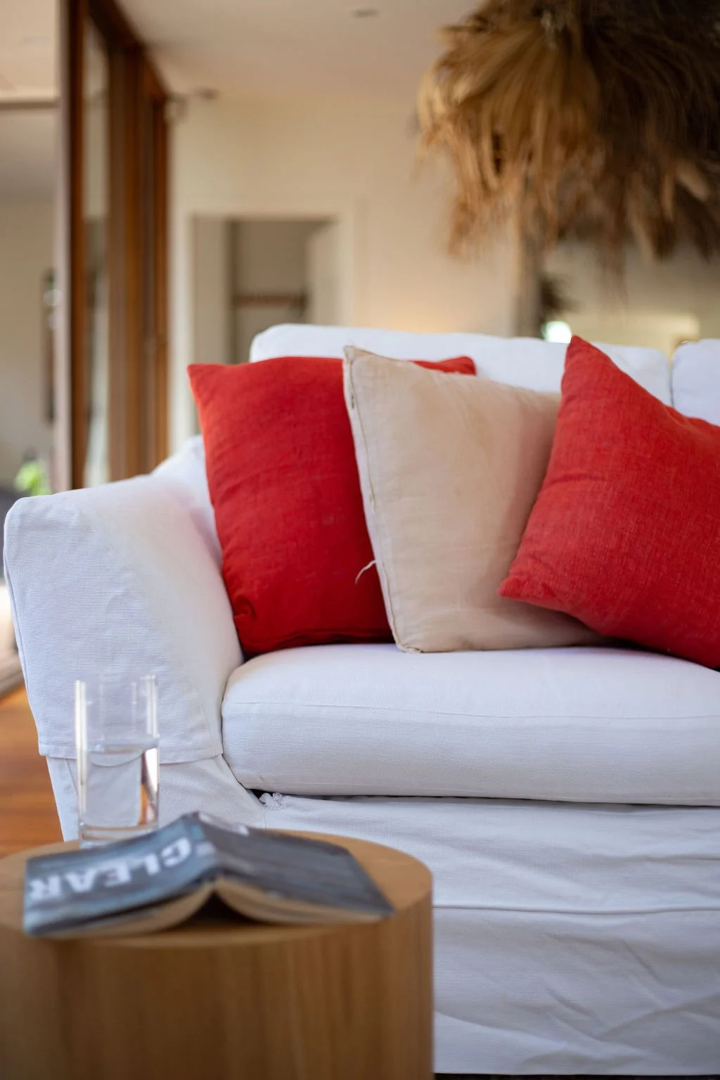 Close-up of a cozy living room scene with a white sofa decorated with red and beige throw pillows, a glass of water on a small wooden side table, and a magazine. The background features a mirror and a tropical wall hanging made of dried palm leaves.