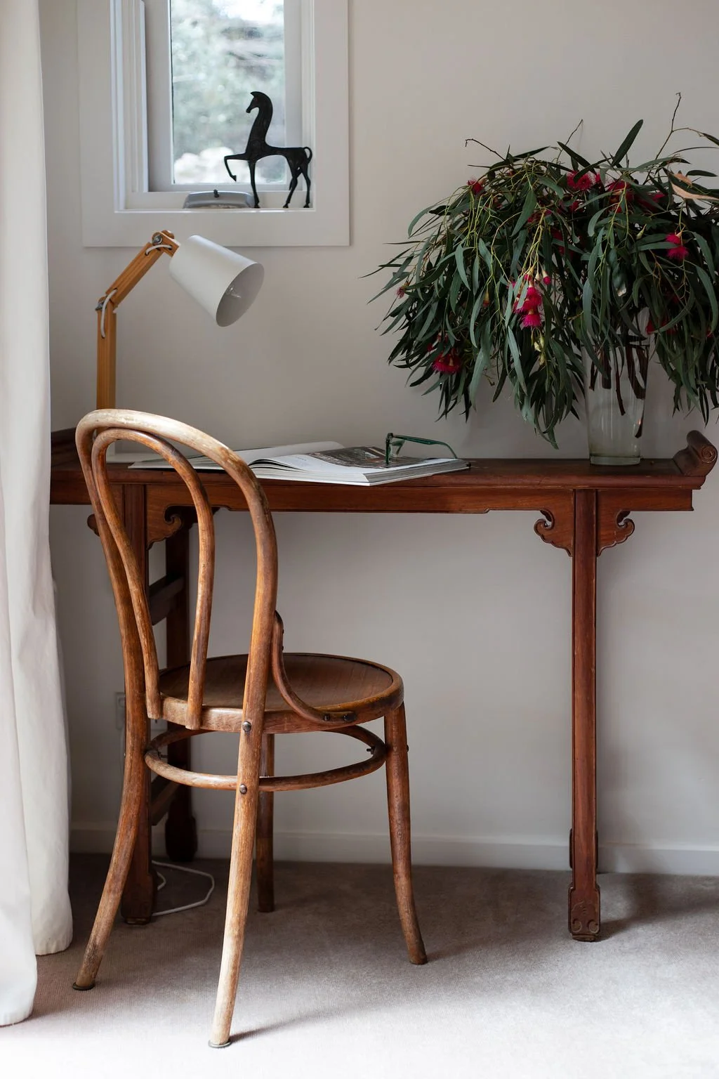 A wooden desk with a large plant in a glass vase, an open magazine on top, and a white adjustable desk lamp. A wooden chair is in front of the desk, and a window with a decorative metal horse sculpture on the sill is in the background.
