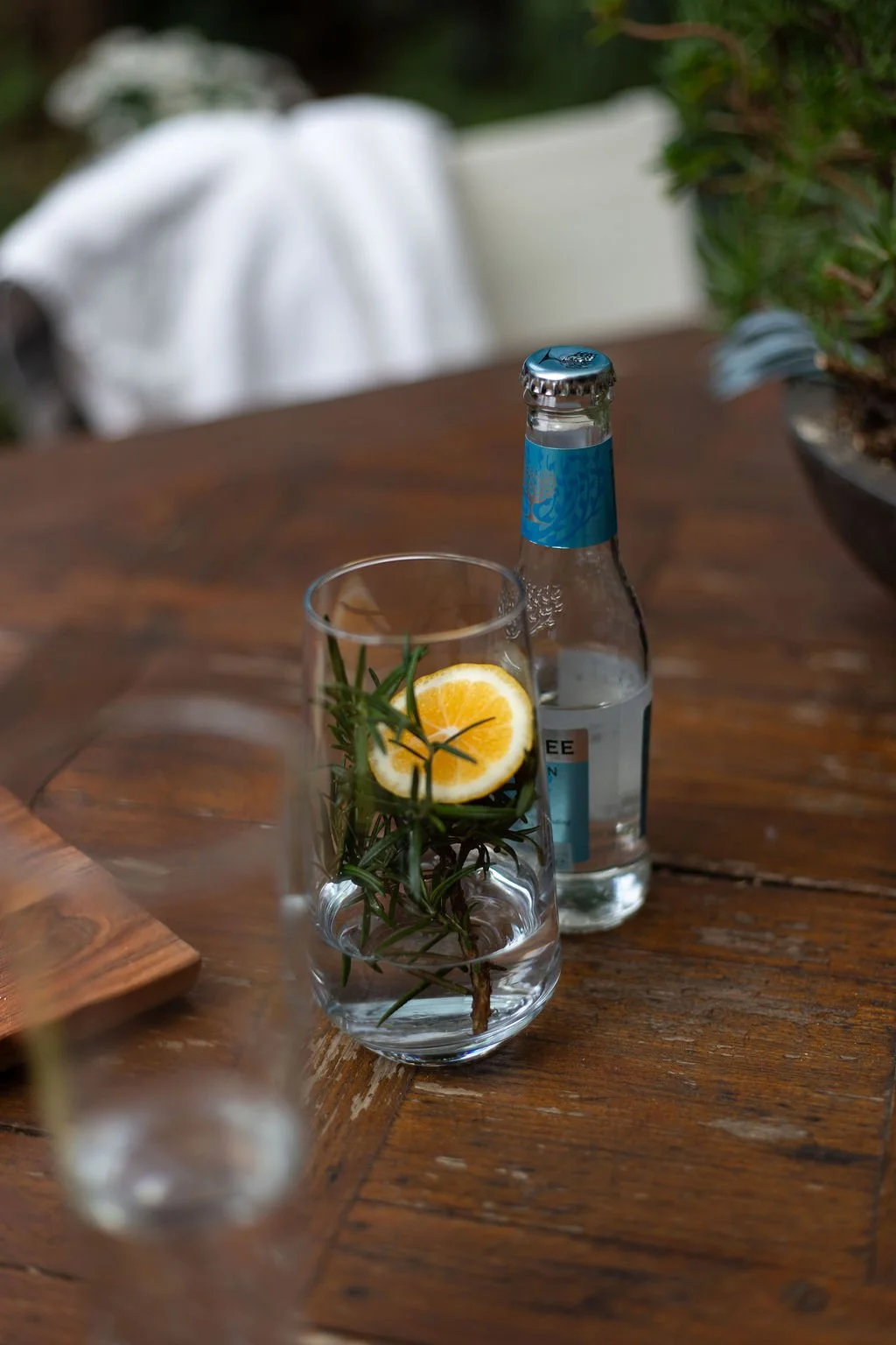 A glass of water with a slice of lemon and fresh rosemary inside, placed on a wooden table next to a bottle of sparkling water.