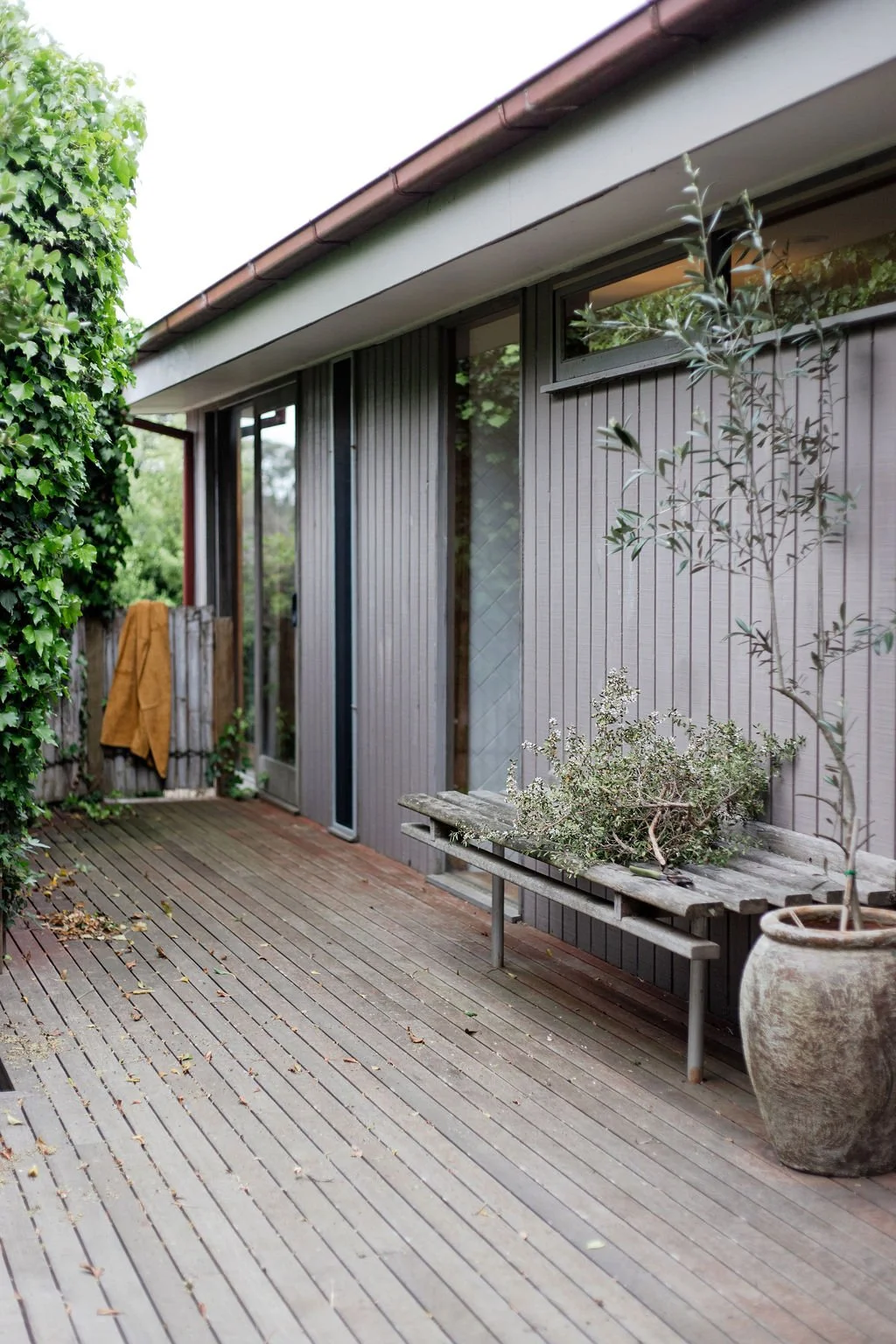 A cozy outdoor wooden deck with plants, a bench, and a house wall with windows.