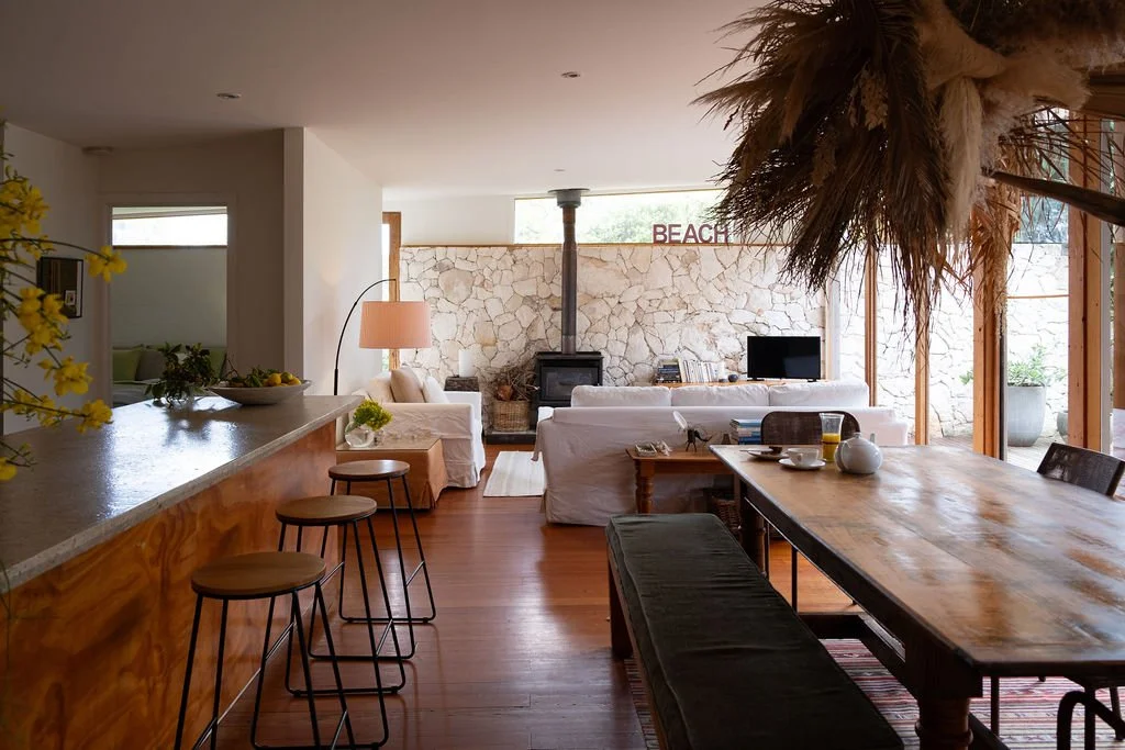 Open-concept living and dining area with wooden floors, stone wall, white sofas, a dining table with chairs, a kitchen counter with bar stools, and large windows with outdoor views.