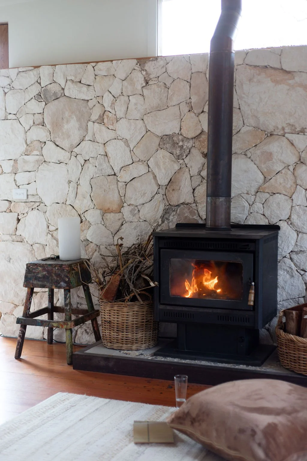 A cozy living room corner with a stone wall, a wood stove with a fire burning, a small rustic stool with a candle, a basket of firewood, and a pillow on a rug.