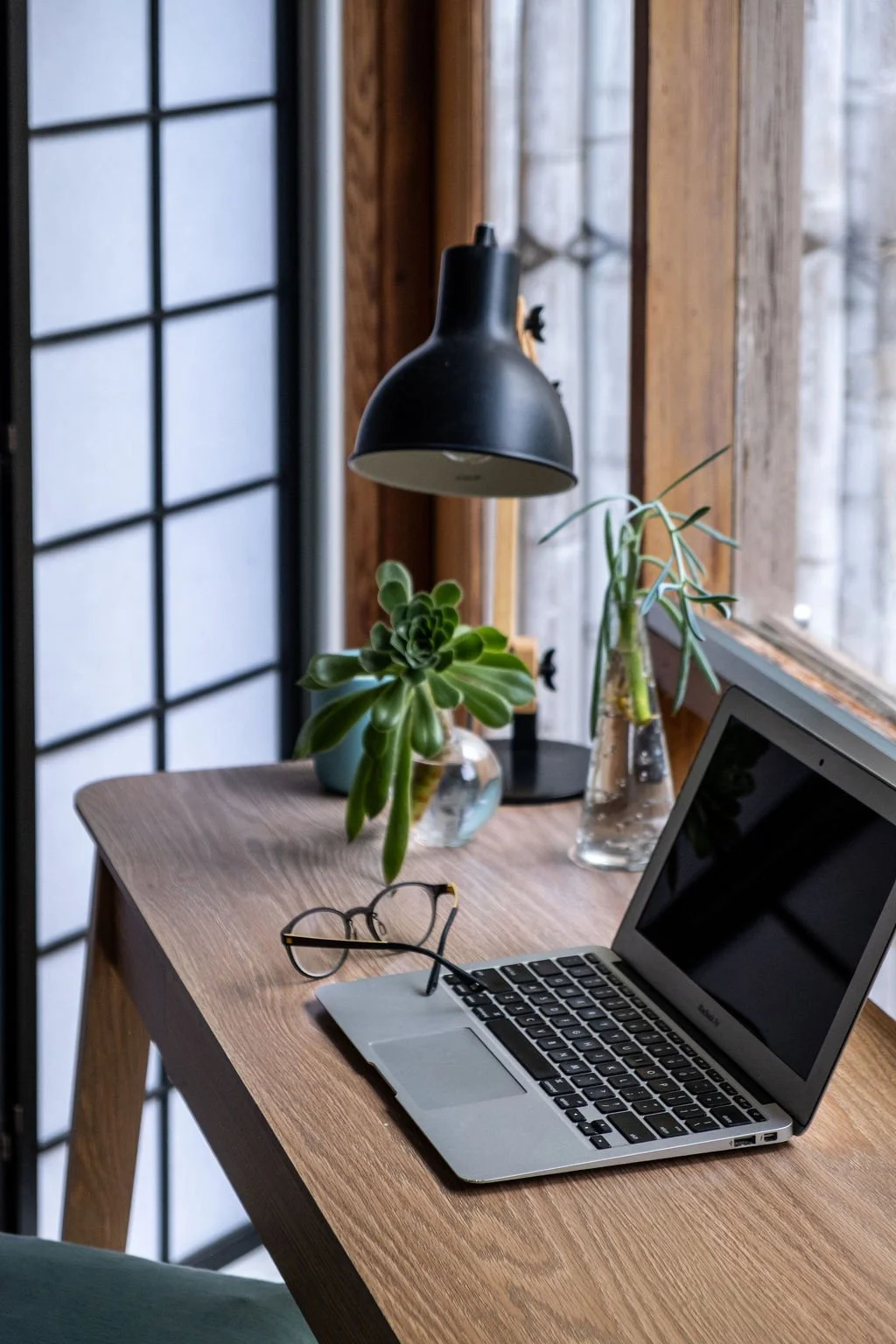 A wooden desk near a window with a laptop, glasses, potted plants, and a black desk lamp.