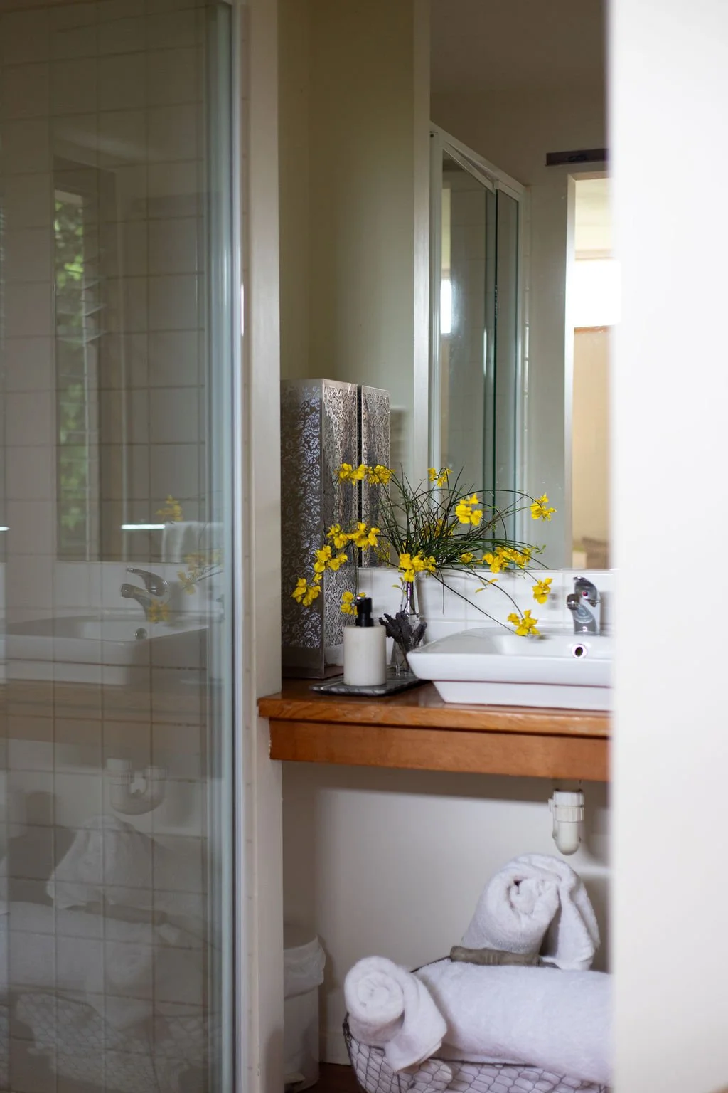 Bathroom with a sink, a mirror, and a shower. Decorative yellow flowers in a vase on the counter. Towels rolled and placed in a basket underneath the counter.