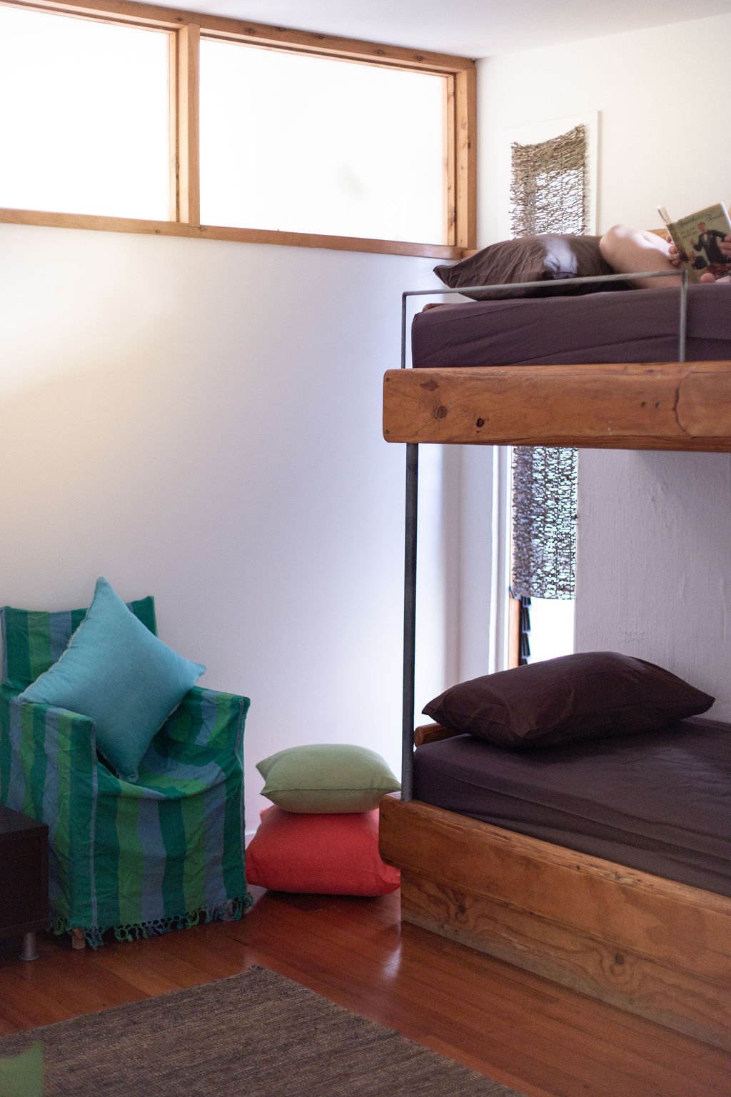 A cozy room with a wooden bunk bed, dark-colored bedding, a green armchair with teal and green cushions, and a small window with natural light.