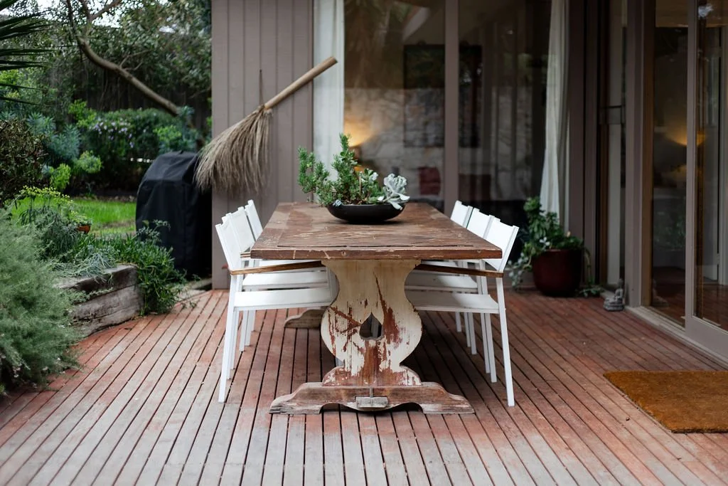 Outdoor wooden deck with a rustic dining table, white chairs, and a black bowl with greenery as a centerpiece.