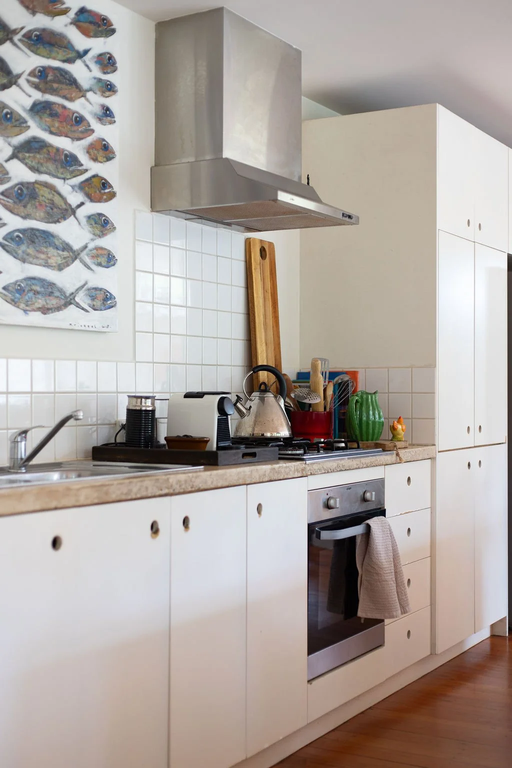 Kitchen with white cabinets, a beige countertop, a stainless steel oven, a stove with a kettle, a white tiled backsplash, a wall art of colorful fish, and various kitchen utensils in a red container and green pottery.
