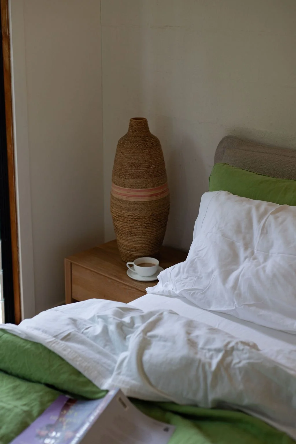 A bedroom with a bed, a green pillow, a white pillow, and a blanket. Next to the bed is a wooden nightstand with a large woven vase, a teacup, and a saucer.