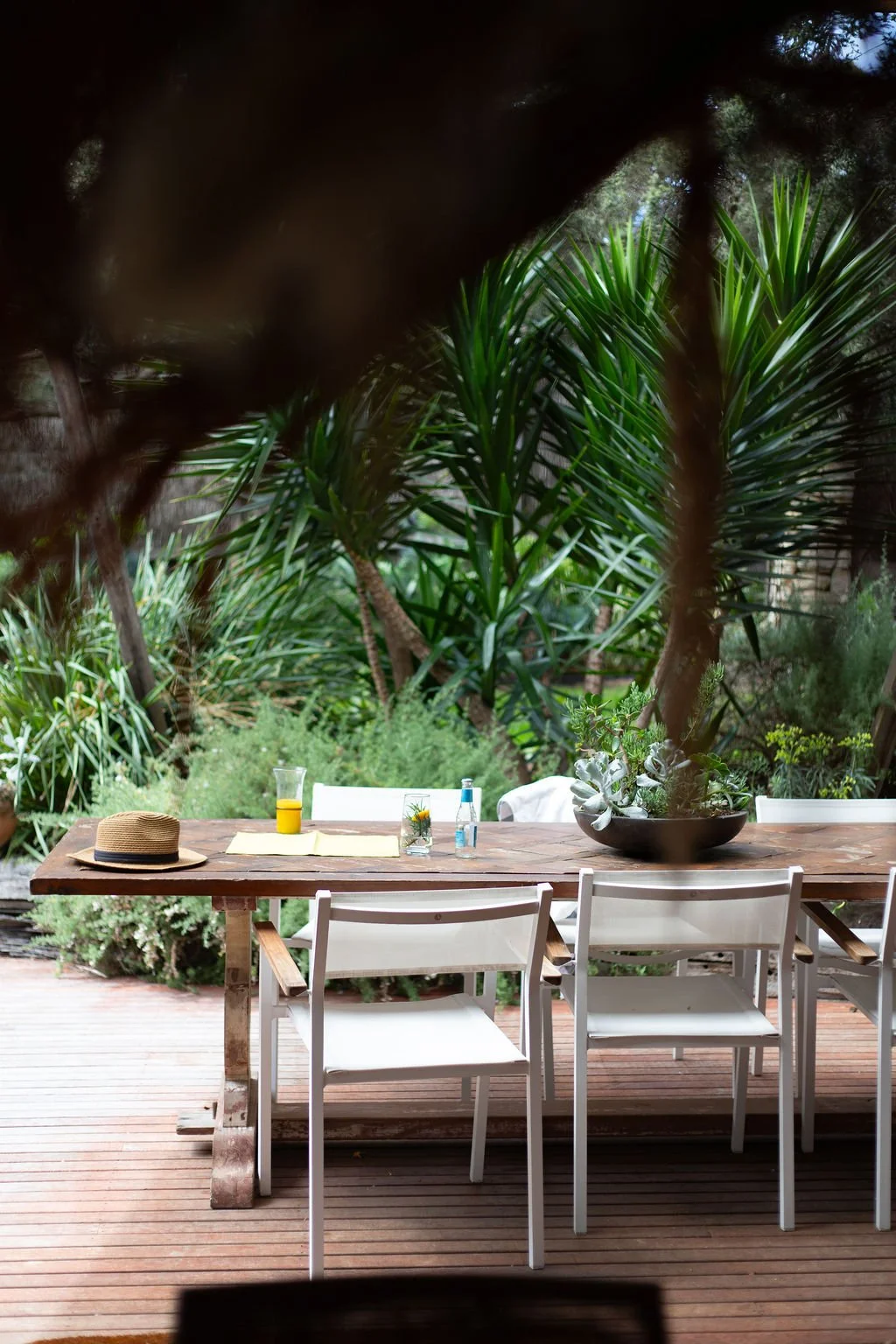 A wooden outdoor table set on a deck with white chairs, decorated with a hat, a glass of orange juice, a small glass bottle, and a black bowl with plants. The background features lush green tropical plants and trees.