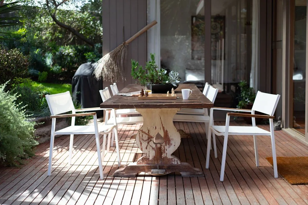 Outdoor patio with a wooden table and four white chairs, plants, and a broom leaning against the wall.