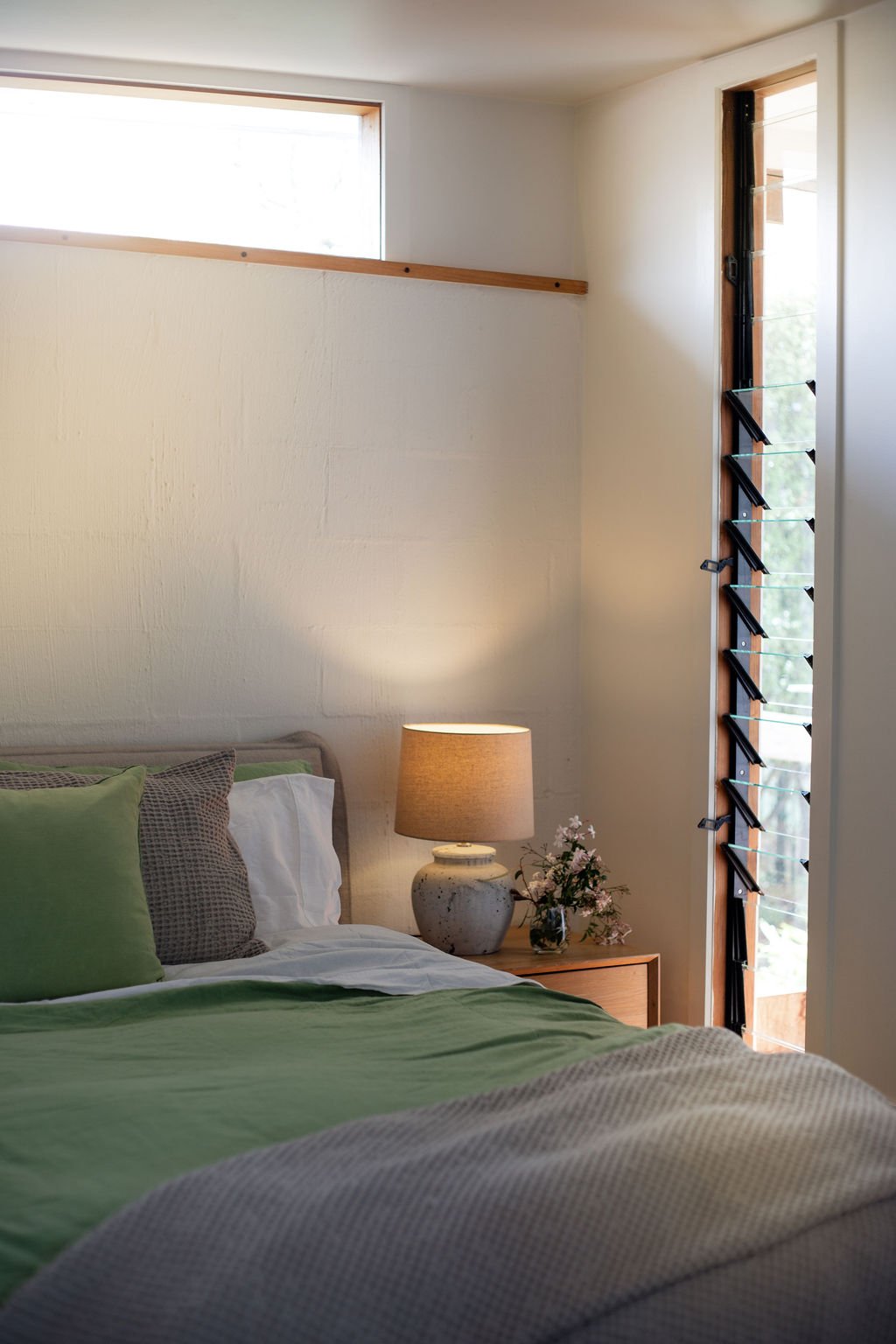 A bedroom with a bed covered in green and white bedding, a bedside table with a lamp and flowers, a window with wooden and metal framing, and sunlight filtering through.