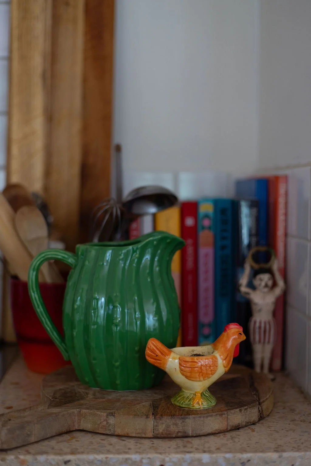 Close-up of a green ceramic pitcher and a small decorative ceramic chicken on a wooden surface in a kitchen, with cookbooks and kitchen utensils in the background.