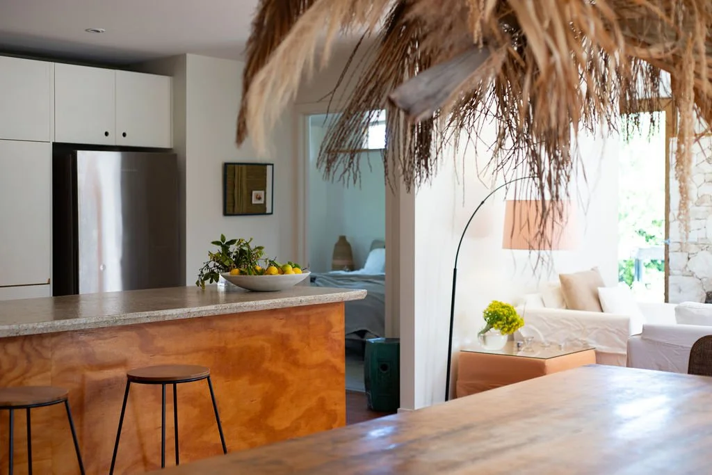 Interior view of a bright living room and kitchen area with a straw ceiling feature, thatched canopy, white couches, a wooden table, a bowl of lemons on the counter, and a bedroom visible in the background.