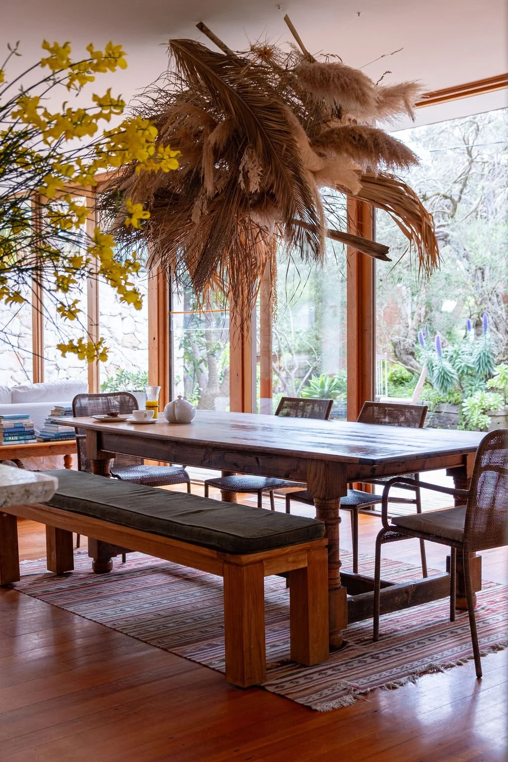 A dining room with a wooden table, chairs, and a bench, decorated with a large hanging arrangement of dried grasses and leaves, in front of large windows revealing a garden outside.