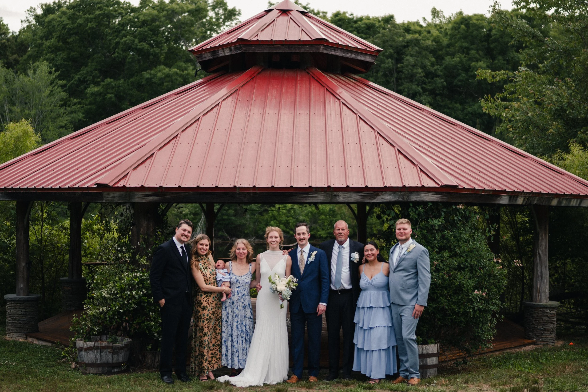 Group of people celebrating with a wedding, standing under a gazebo with a red metal roof, surrounded by greenery.