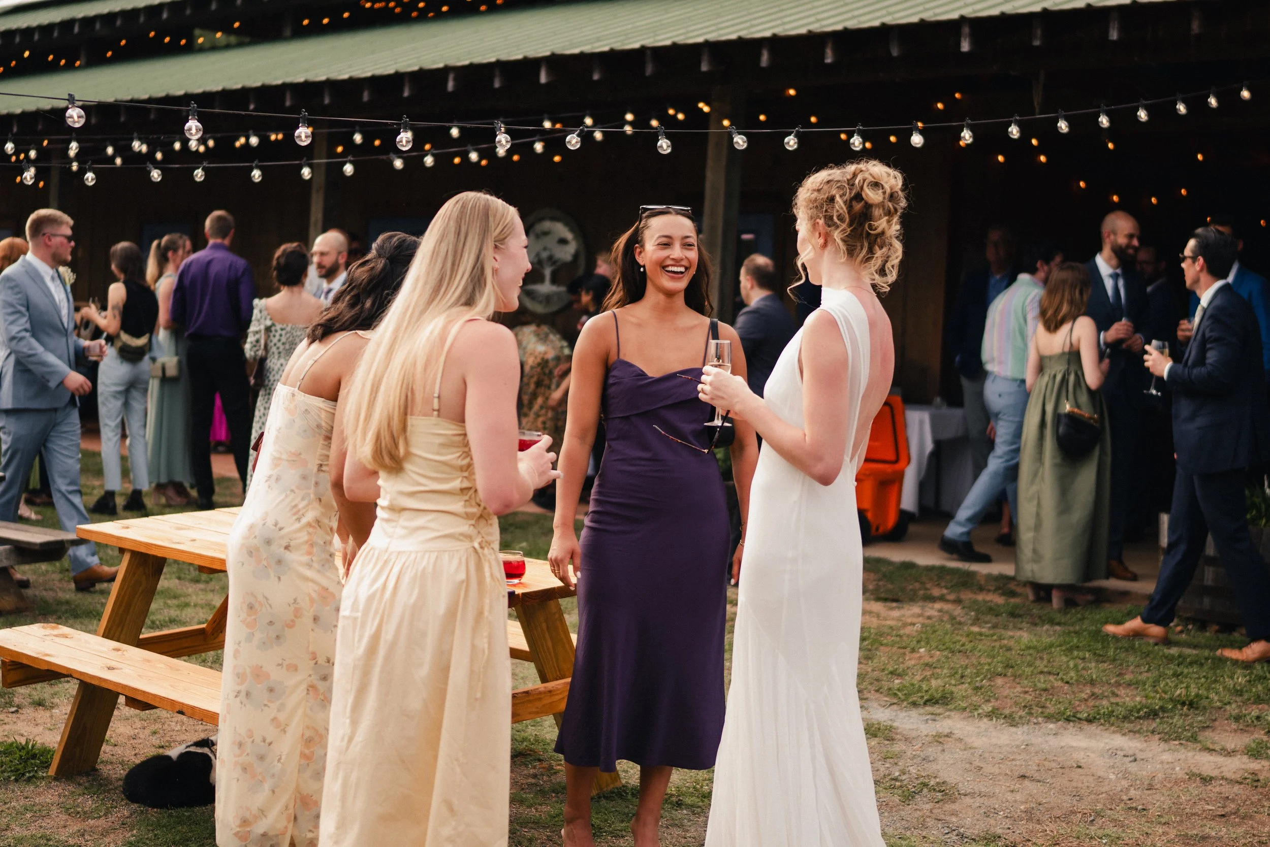 A group of women in dresses talking and laughing at an outdoor gathering, with other people in the background under string lights and a green roofed structure.