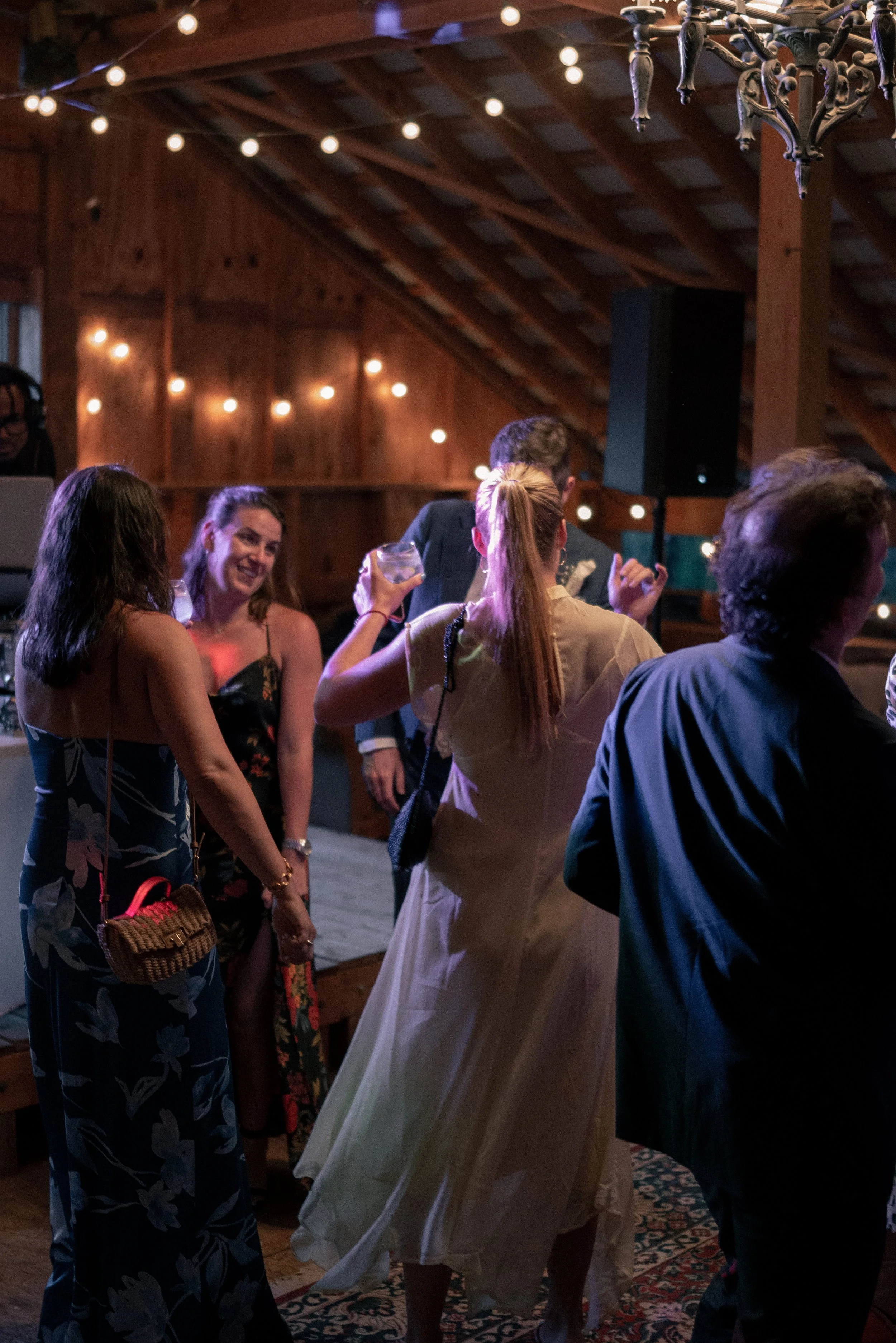 People dancing and socializing at an indoor party or celebration in a wooden venue with string lights and a chandelier.