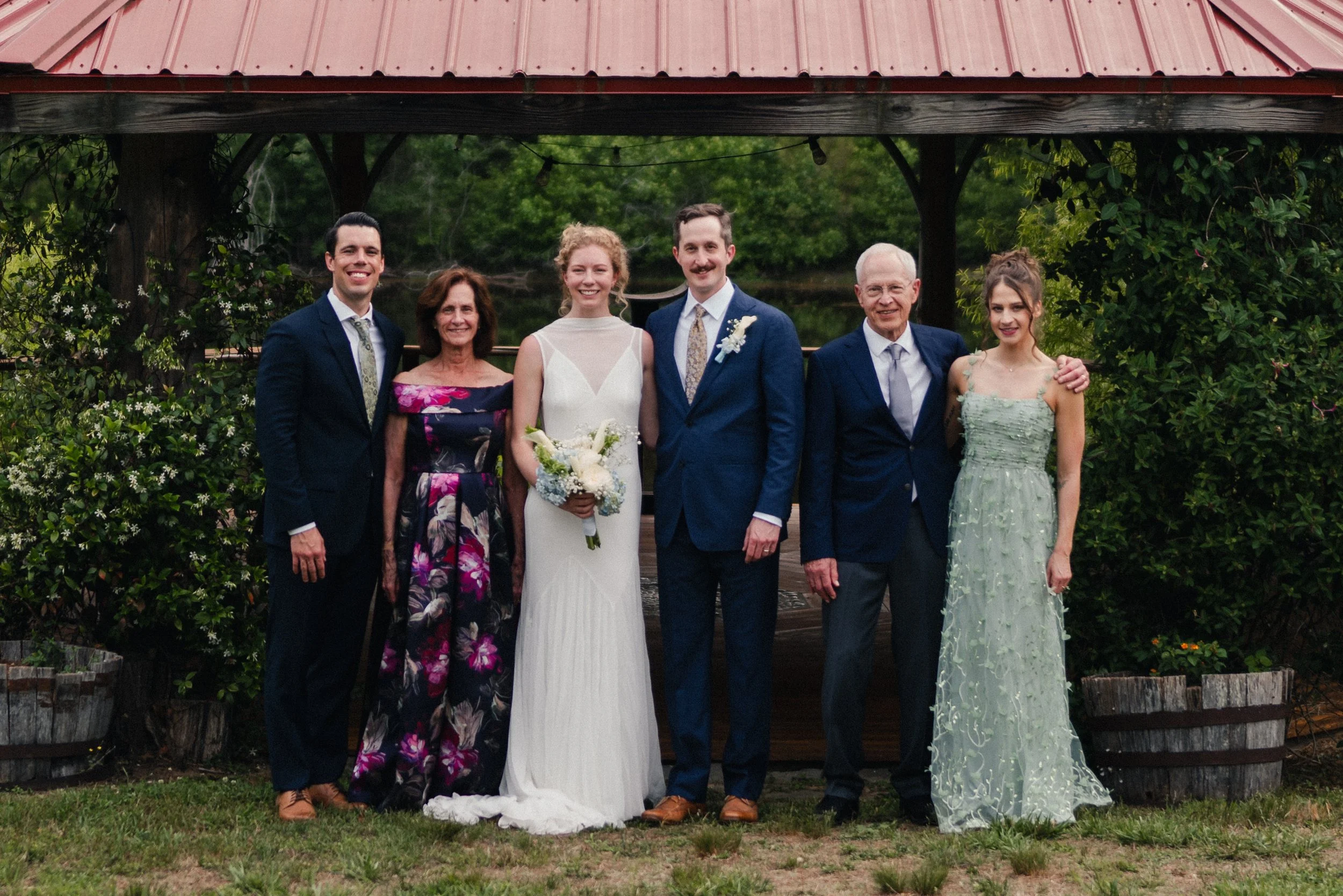 Group of seven people, including a bride and groom, posing outdoors near a wooden gazebo and greenery, celebrating a wedding.