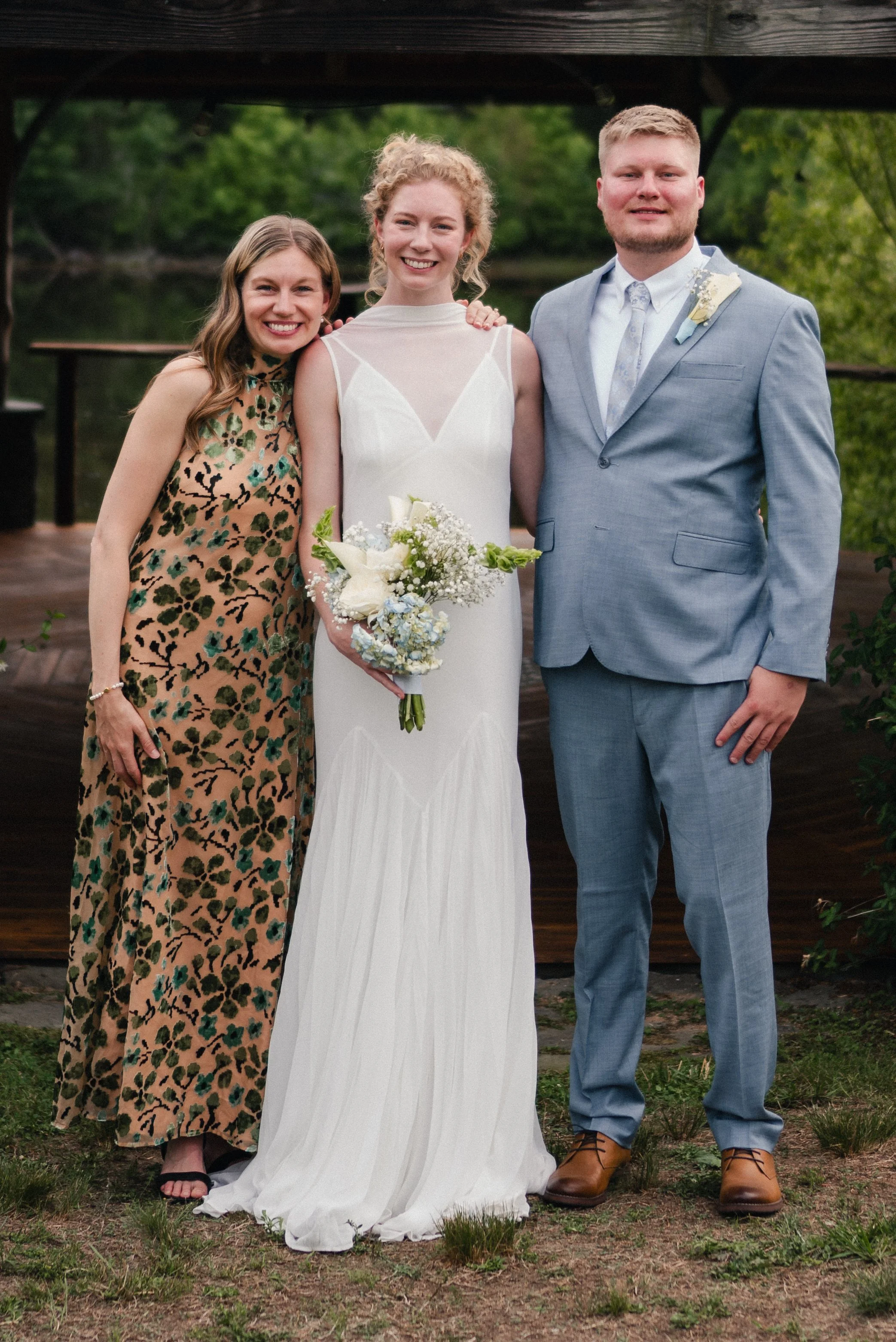 Group of three people standing outdoors, smiling for a photo, with greenery and a wooden structure in the background. The woman in the center is wearing a white wedding dress holding a bouquet, flanked by a woman in a animal print dress and a man in 