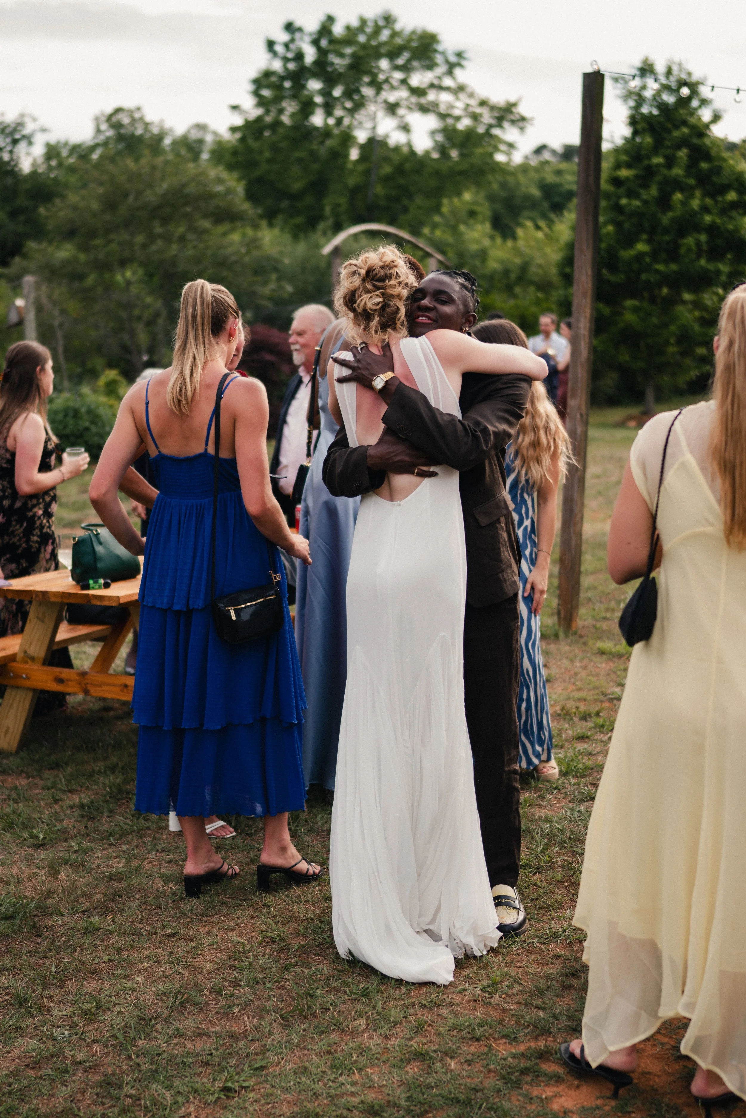 A group of people at an outdoor wedding reception, with a couple hugging in the center. The bride is wearing a white gown and the groom is in a dark suit. Other guests are standing around, talking and socializing, with trees and string lights in the 
