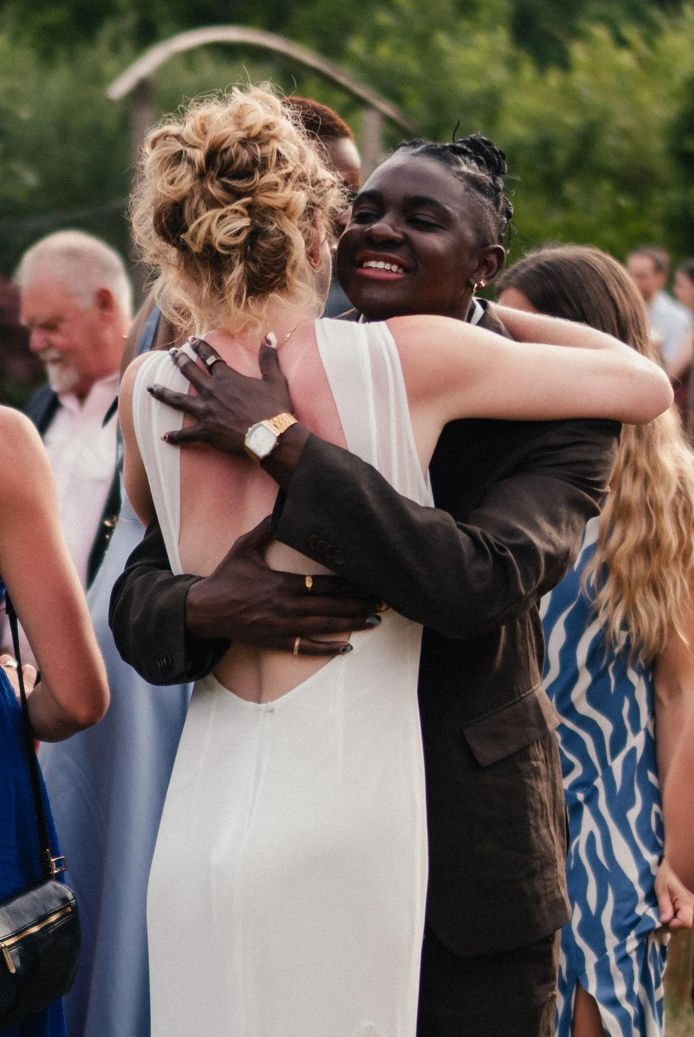 Two women embrace at an outdoor gathering, smiling and hugging among other people.
