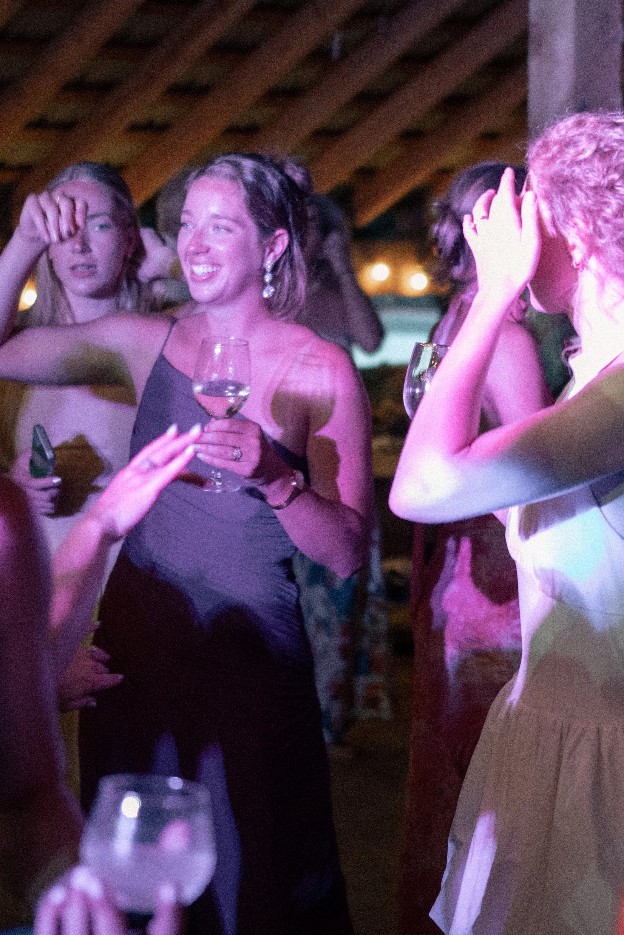 People at a social gathering, some with glasses of wine, under a wooden ceiling with purple lighting.