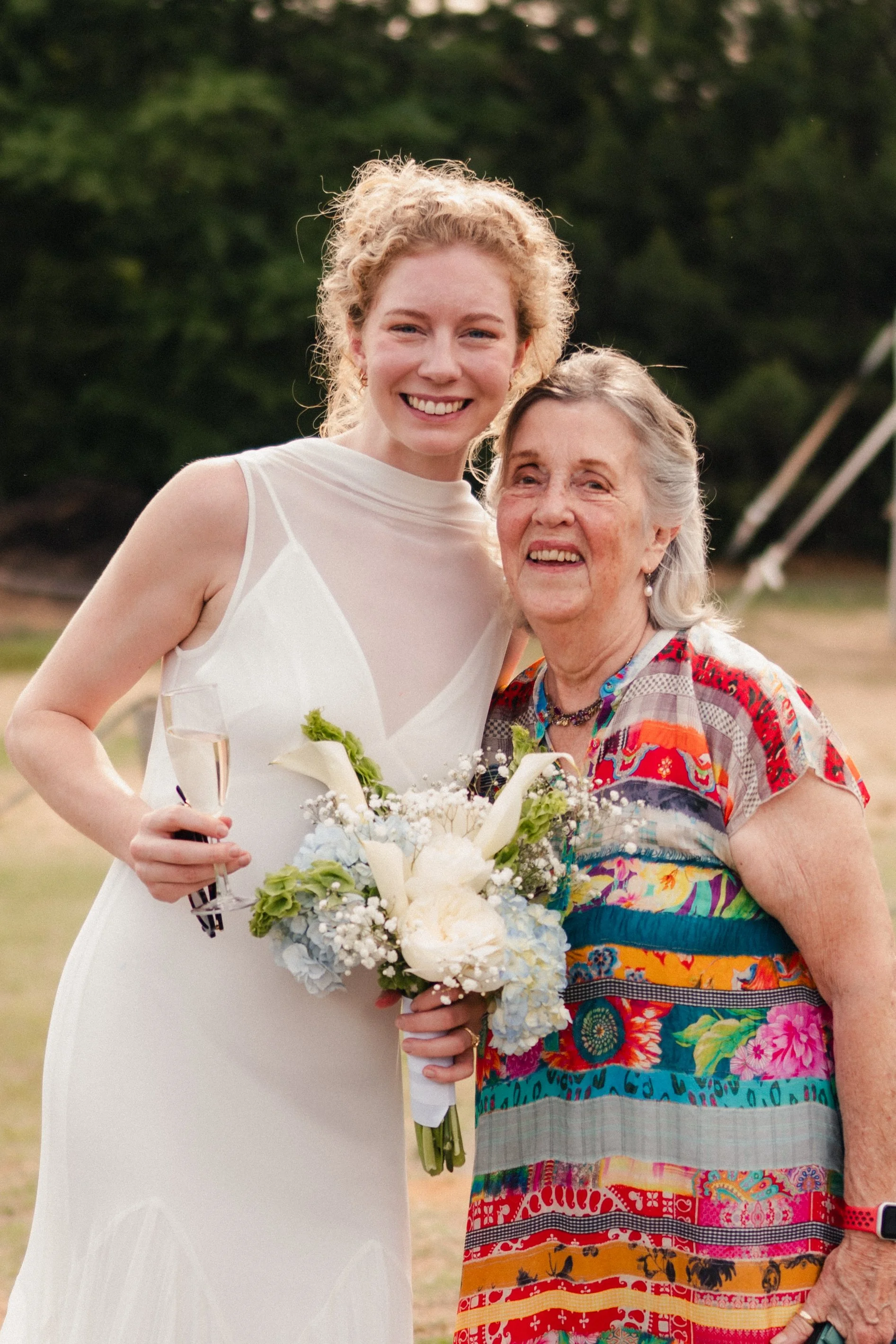 A bride in a white dress holding a bouquet and a champagne glass, standing next to an older woman in a colorful patterned dress outdoors with trees in the background.