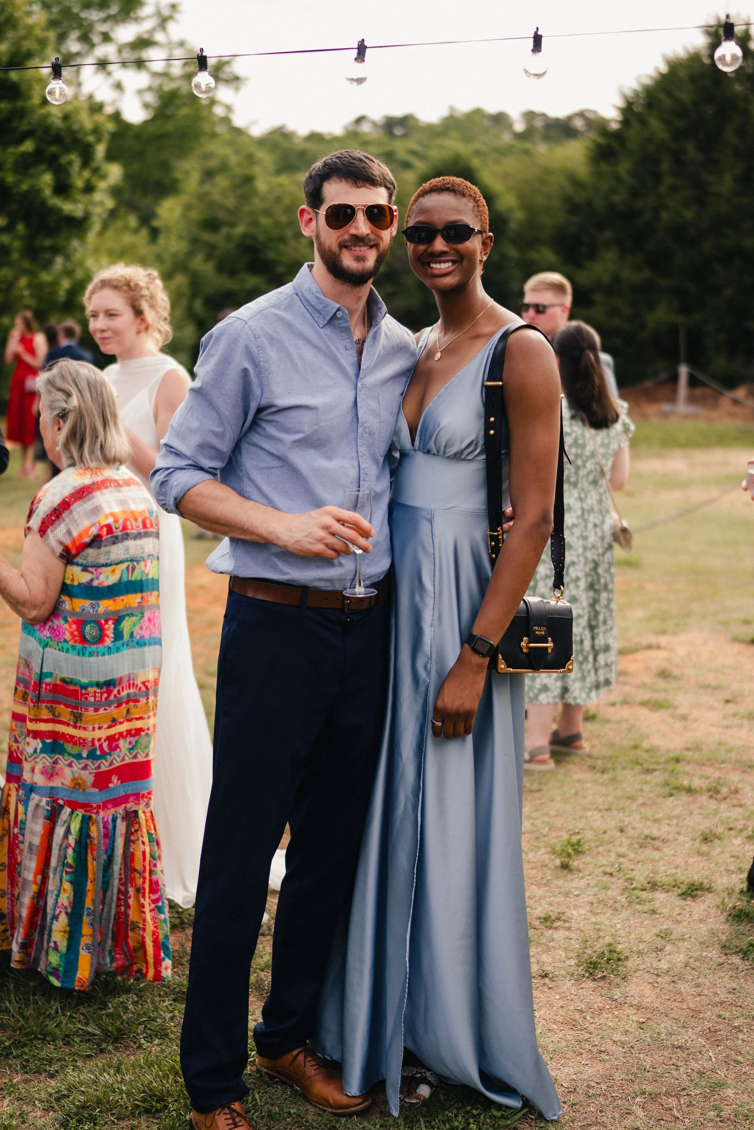 A man and woman wearing sunglasses standing outdoors at a social event, smiling, with string lights hanging overhead and other guests in the background.