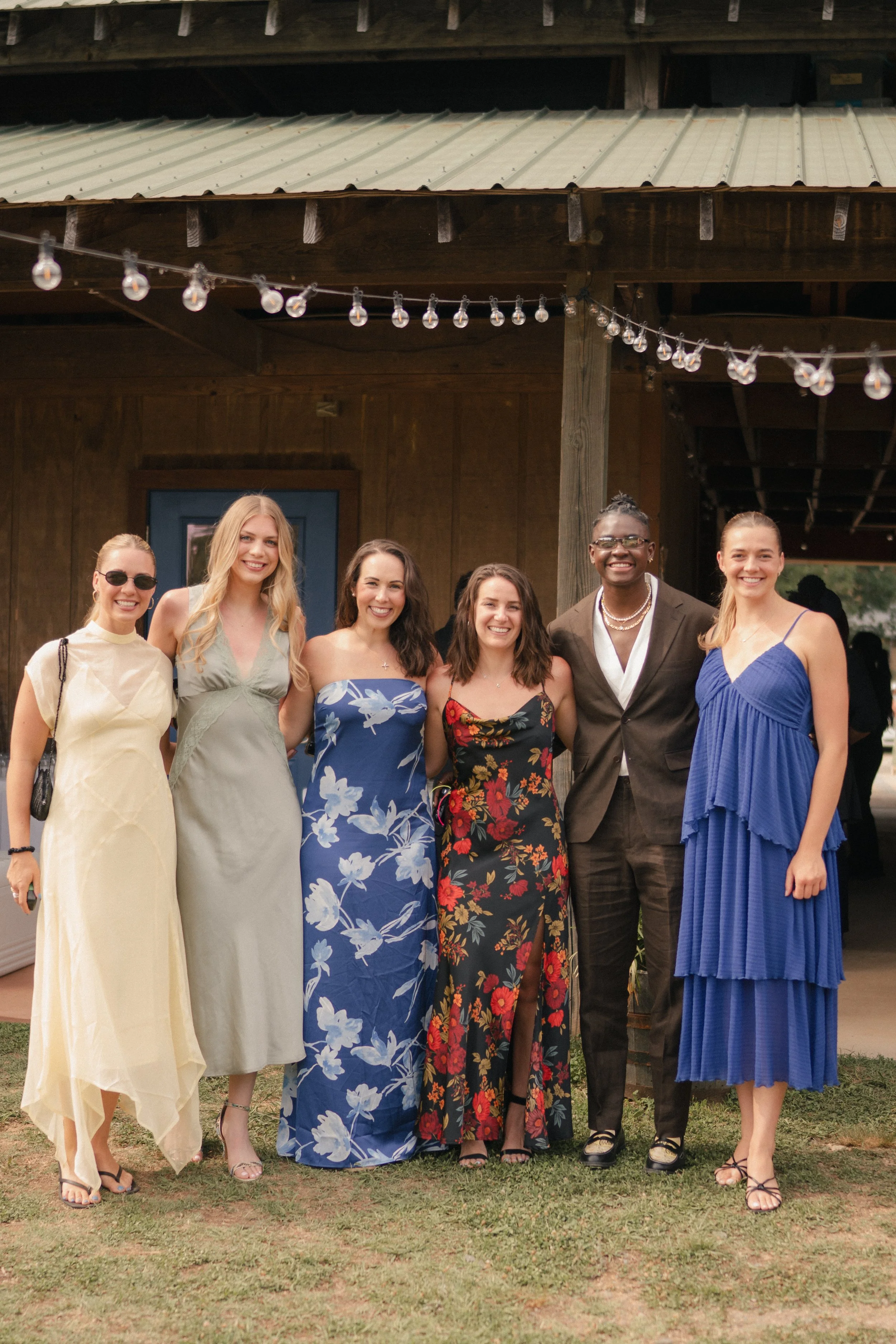 A group of six women and one man standing outdoors in front of a wooden building with string lights hanging above them, all smiling and dressed in formal or semi-formal attire.