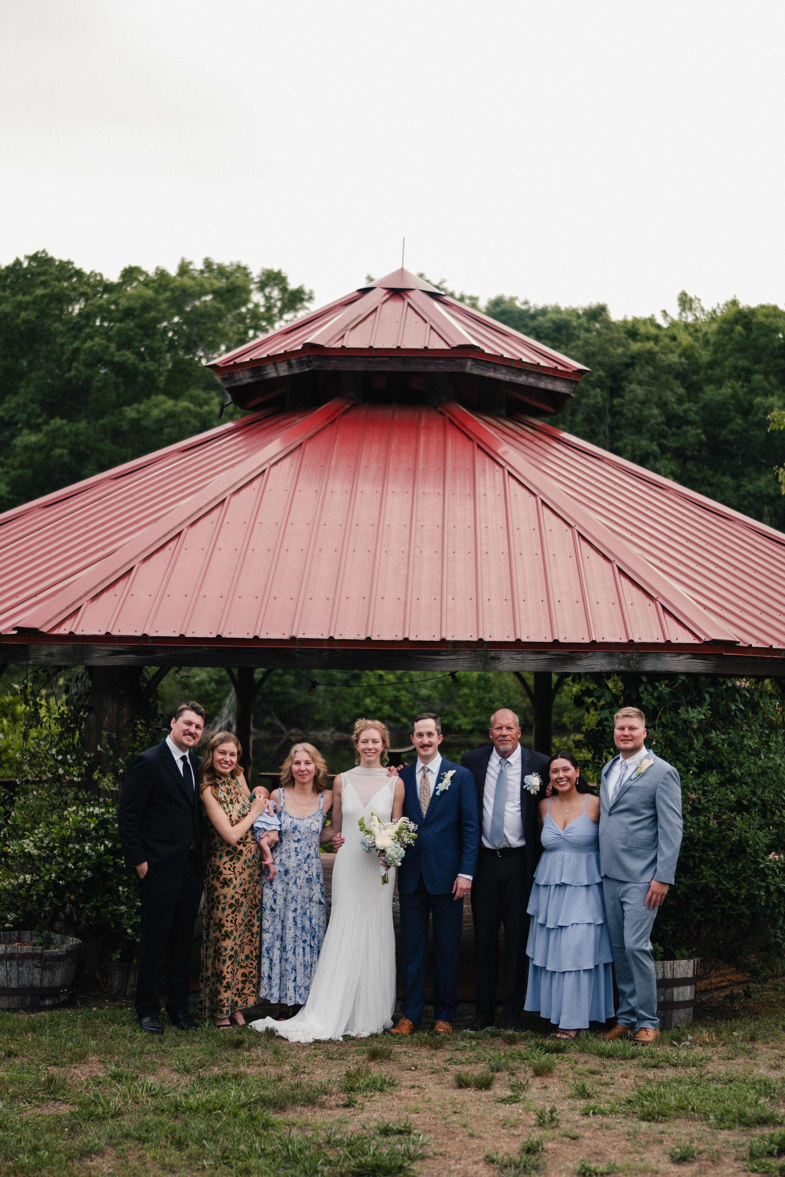 A group of people dressed in formal attire posing outdoors under a pavilion with a red metal roof for a wedding photo, with trees in the background.
