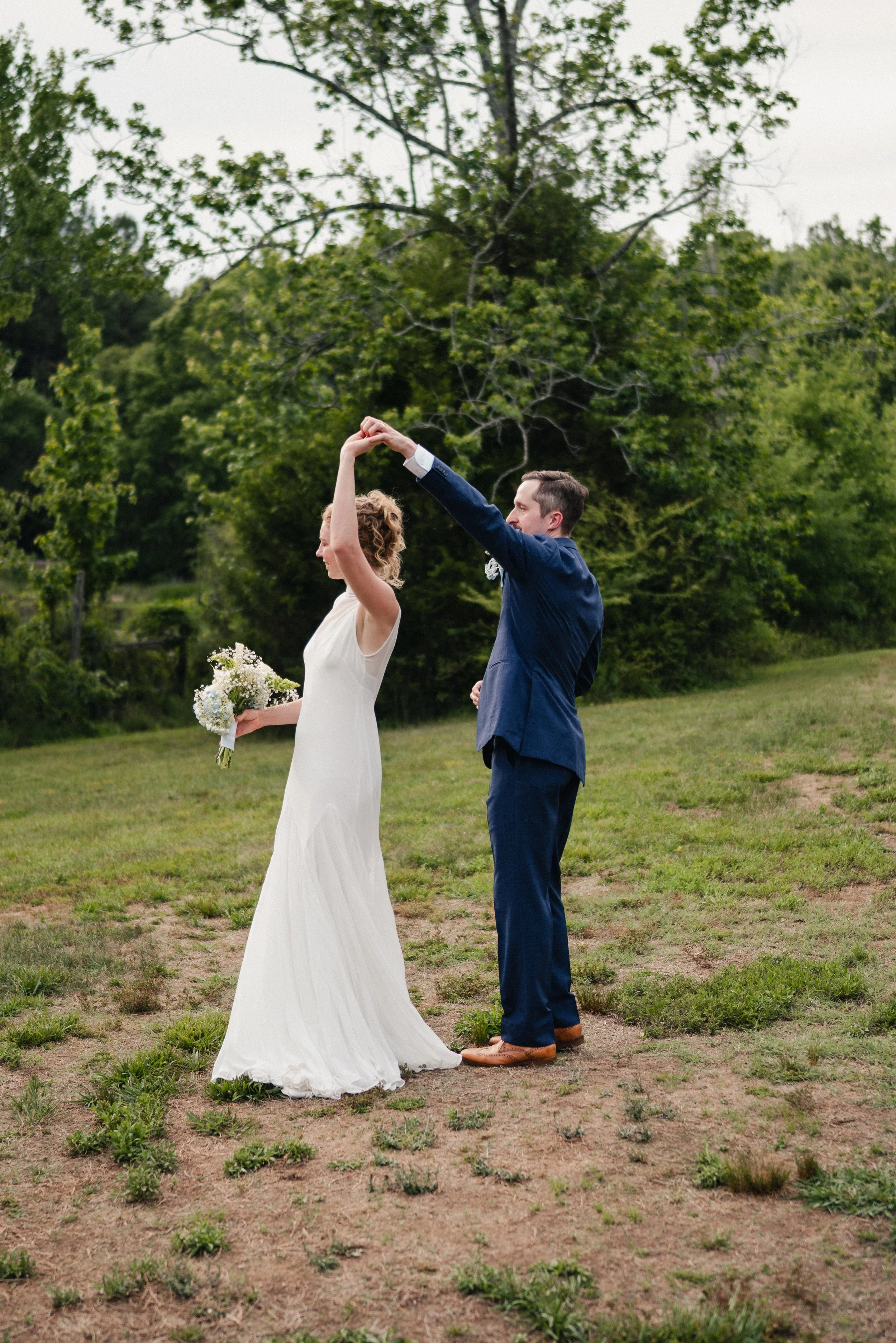 A bride and groom are dancing outside on grass, with the groom twirling the bride. The bride holds a bouquet of flowers and wears a white wedding dress, while the groom is dressed in a blue suit with brown shoes. There are trees and greenery in the b