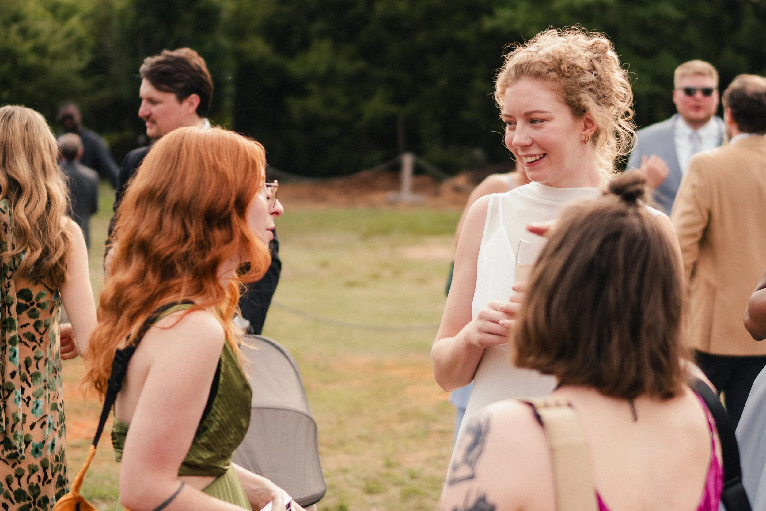 People socializing outdoors, including women with red and blonde hair, and men in formal attire, in a grassy park area with trees in the background.