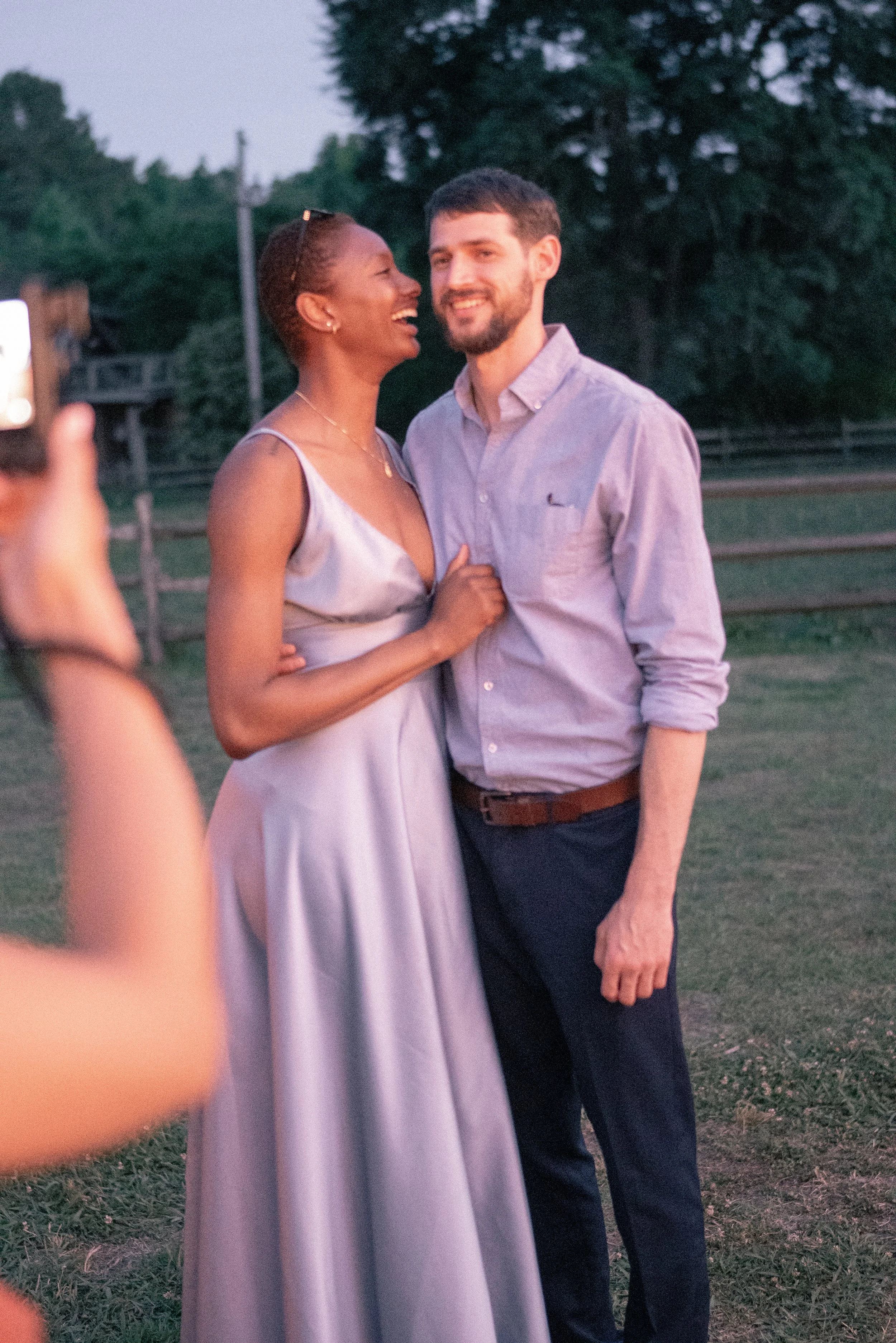 A woman in a light dress and a man in a dress shirt smiling and laughing together outdoors during dusk or evening.