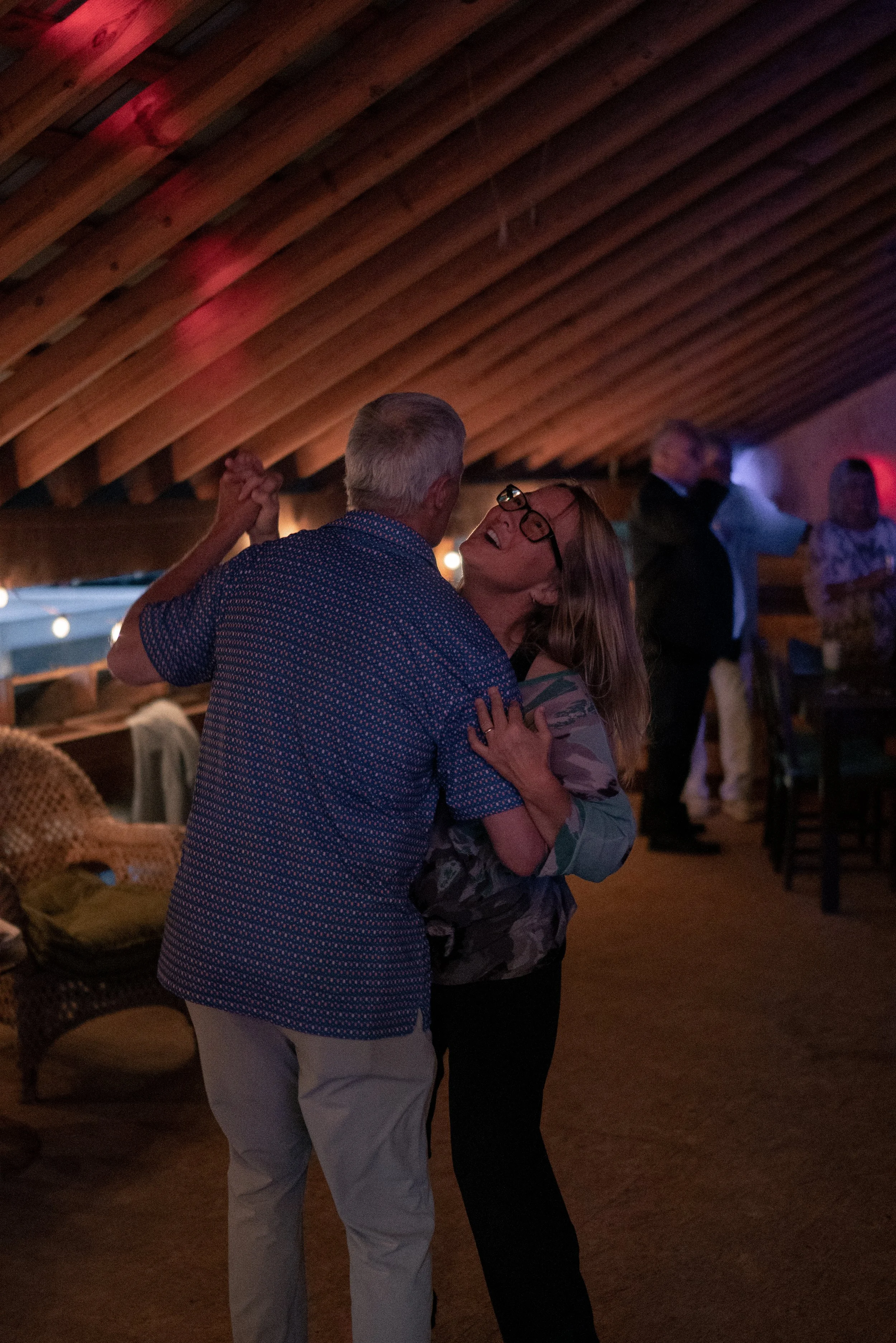 An elderly man and woman dancing closely together in a dimly lit indoor setting with a wooden ceiling, surrounded by other people.